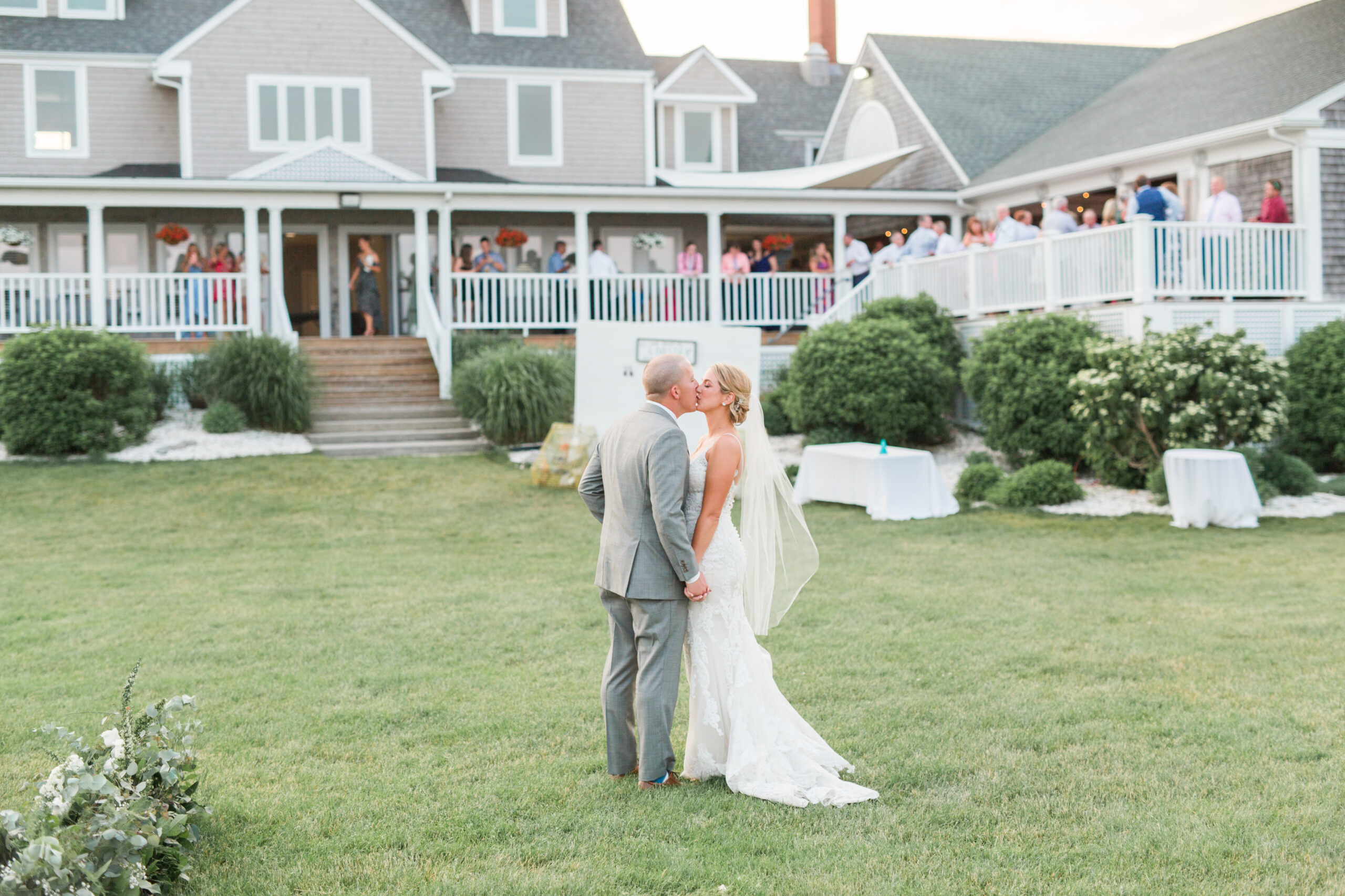 Bride and groom kissing during their wedding cocktail hour with guests on the porch behind them