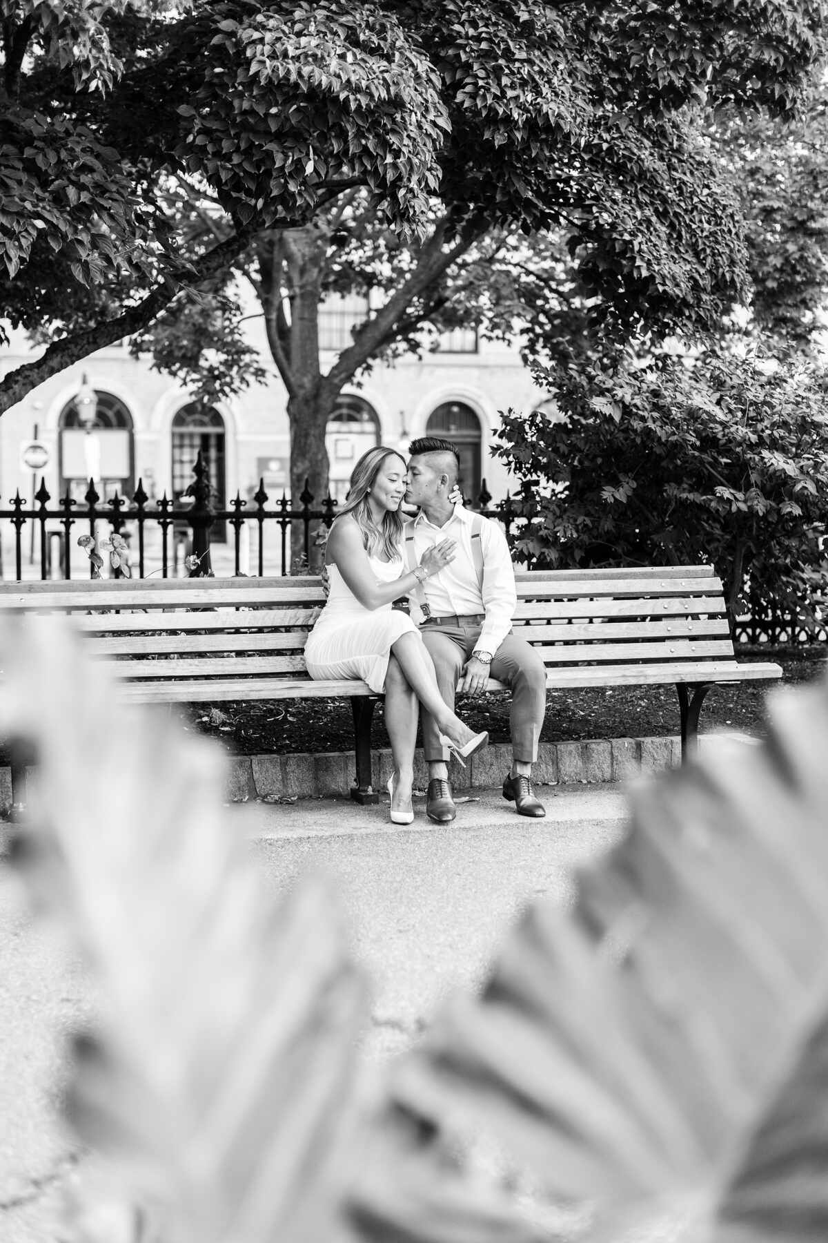 A black and white photo of a couple sitting closely together on a classic wooden park bench at the Boston Public Garden. The man gently kisses the woman on the cheek as she smiles and rests her hand on his chest. Ornate iron fencing, lush trees, and elegant arched Boston architecture are softly blurred in the background, captured by a photographer in Boston.