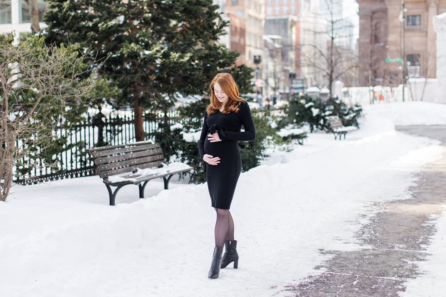 A pregnant woman with red hair stands alone on a snowy path in Boston Public Garden, looking down and cradling her baby bump with both hands. She wears a black ribbed dress, black tights, and ankle boots, with snow-dusted benches and evergreen trees behind her.