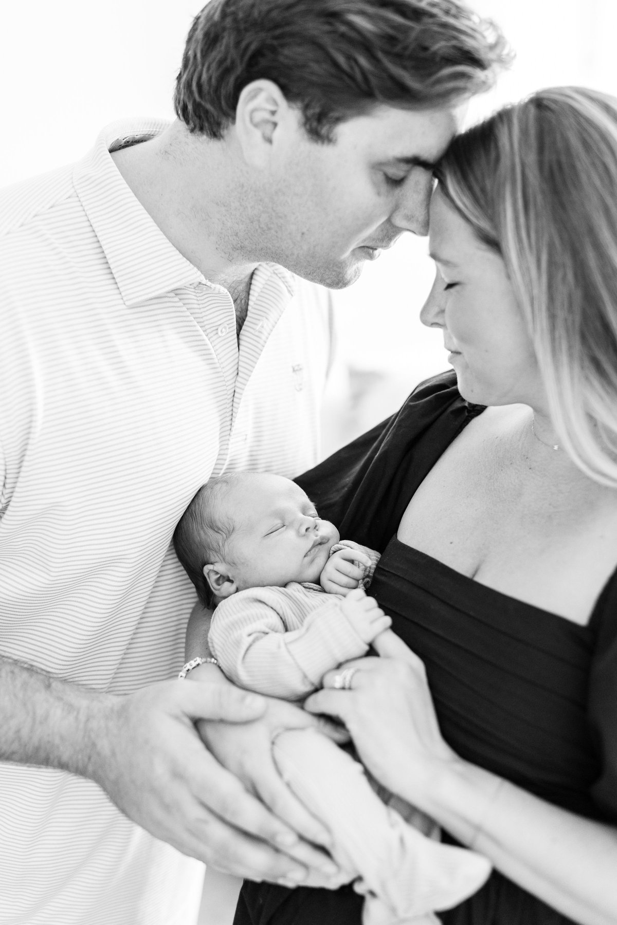 Newborn photo shoot at home close-up in black and white of parents touching foreheads above their sleeping newborn baby who is nestled between them in a ribbed outfit