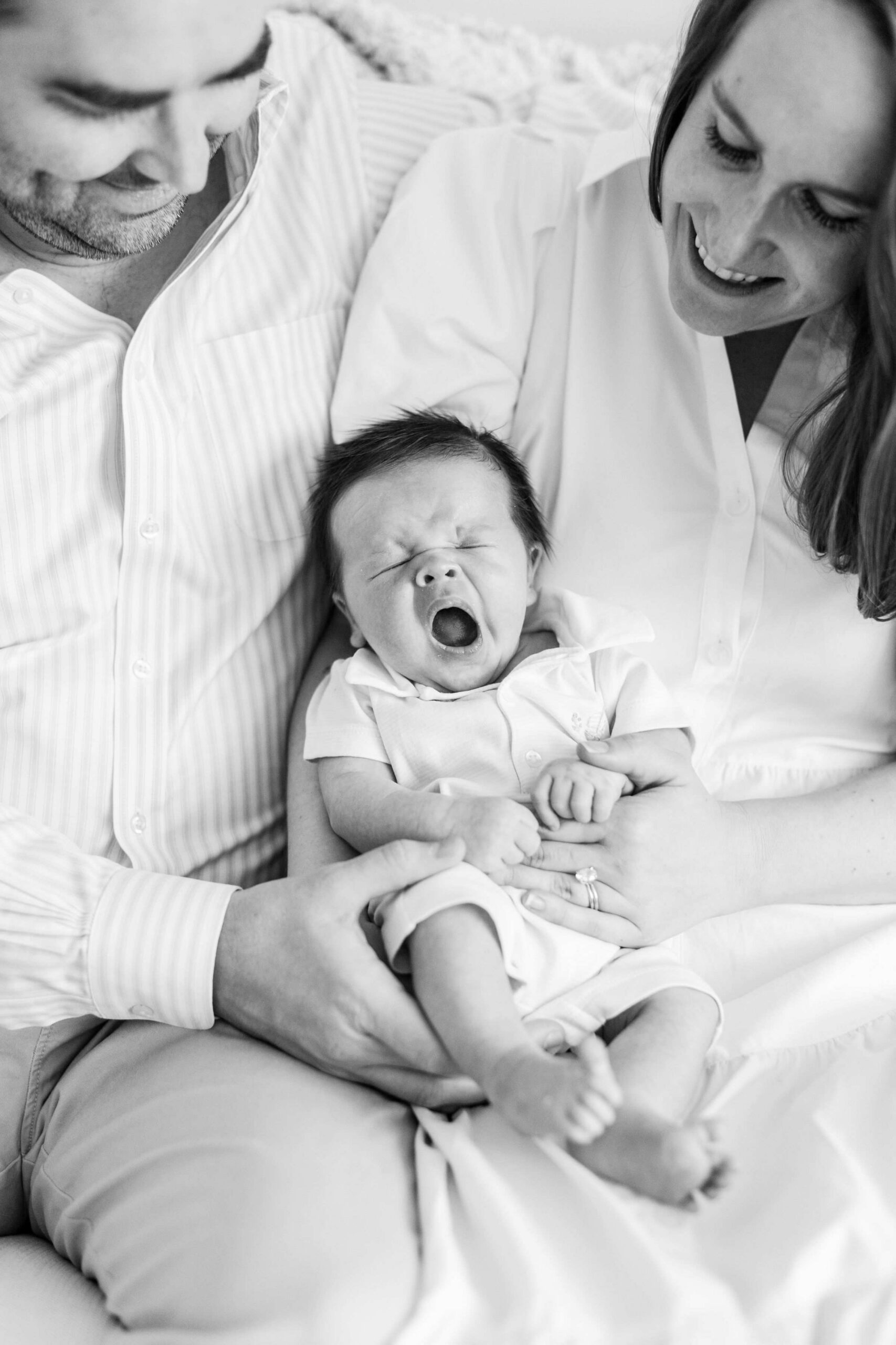 Tips for newborn photo shoot captured in an unposed moment of parents laughing while their newborn baby lets out a big yawn between them in black and white