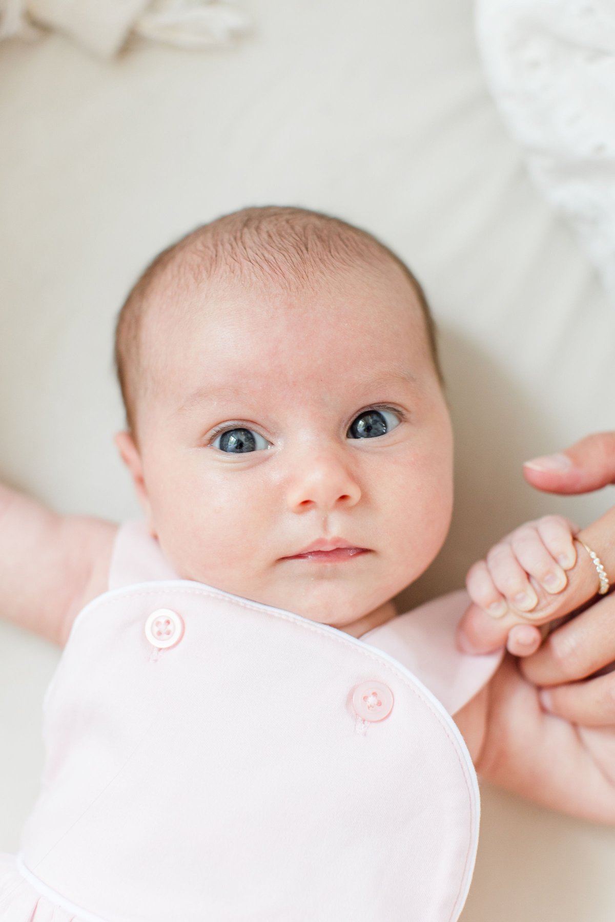 Newborn photos at home close-up of a wide-awake newborn baby girl in a soft pink outfit with striking blue eyes gazing up at the camera while holding a parent's finger