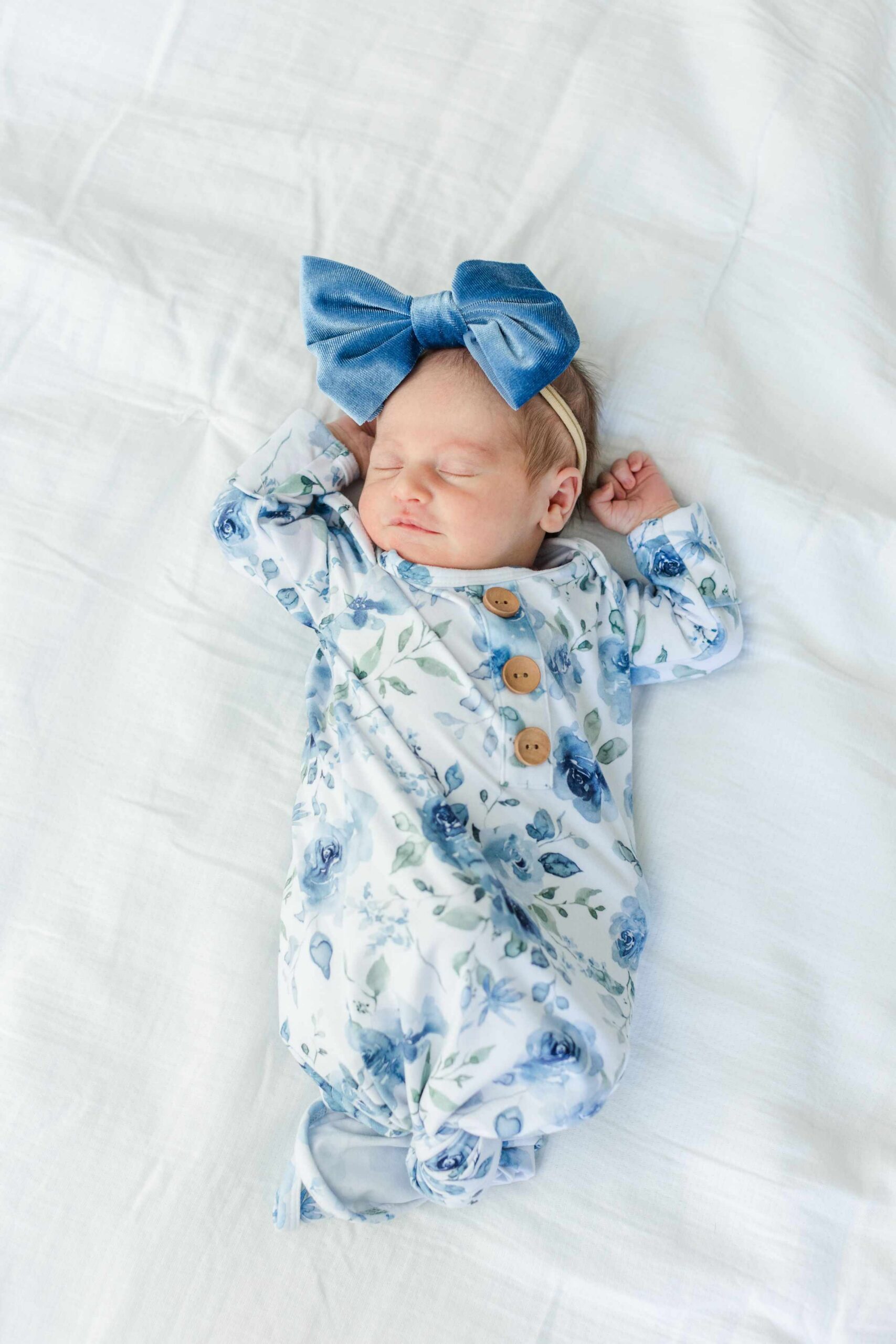 Newborn photos at home of a sleeping baby girl in a blue watercolor floral knotted gown with a large velvet blue bow headband lying on a white bed