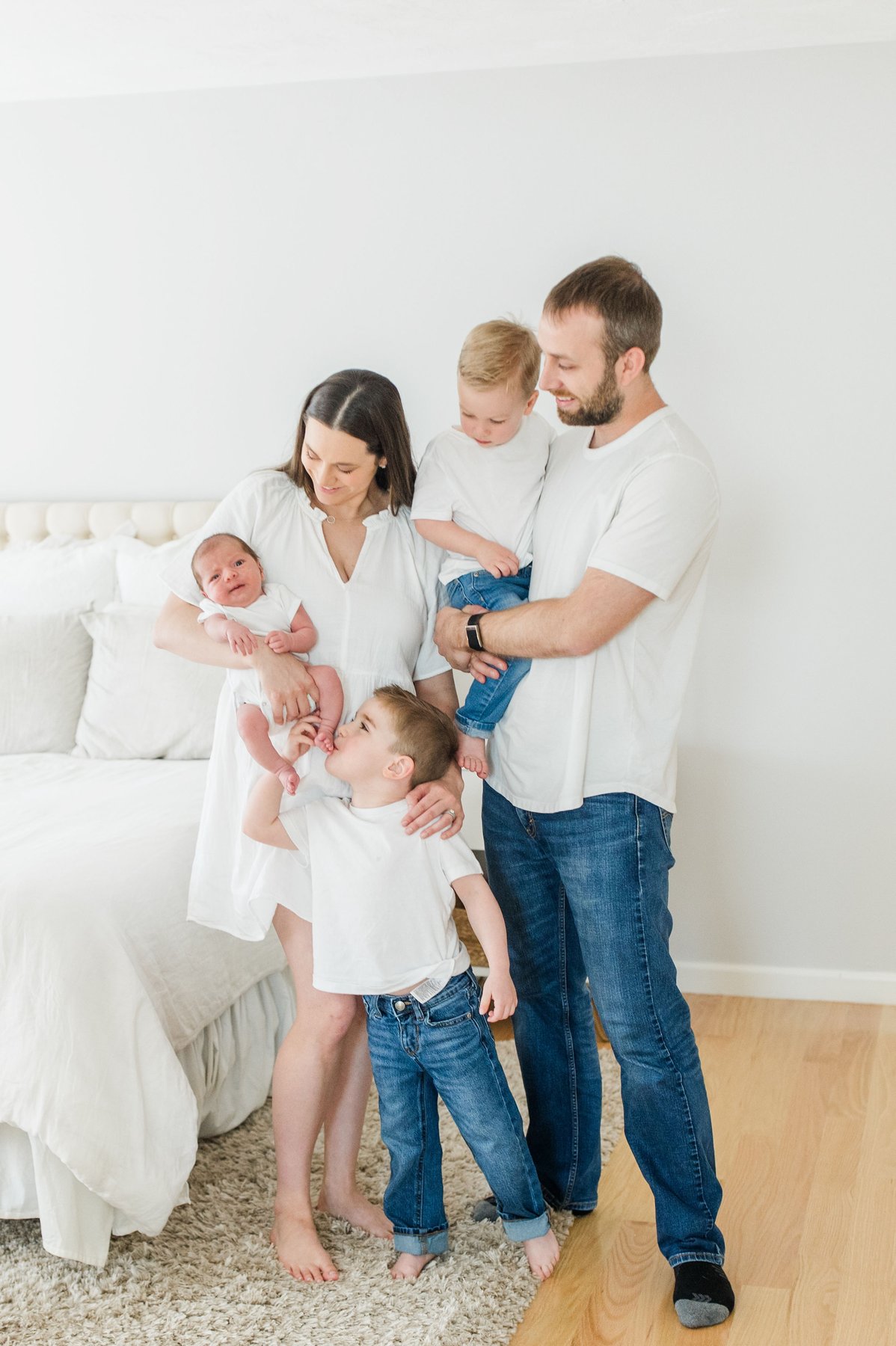 Newborn photos at home of a family of five with mom holding their newborn, dad holding a toddler, and an older boy kissing the baby beside a tufted white bed