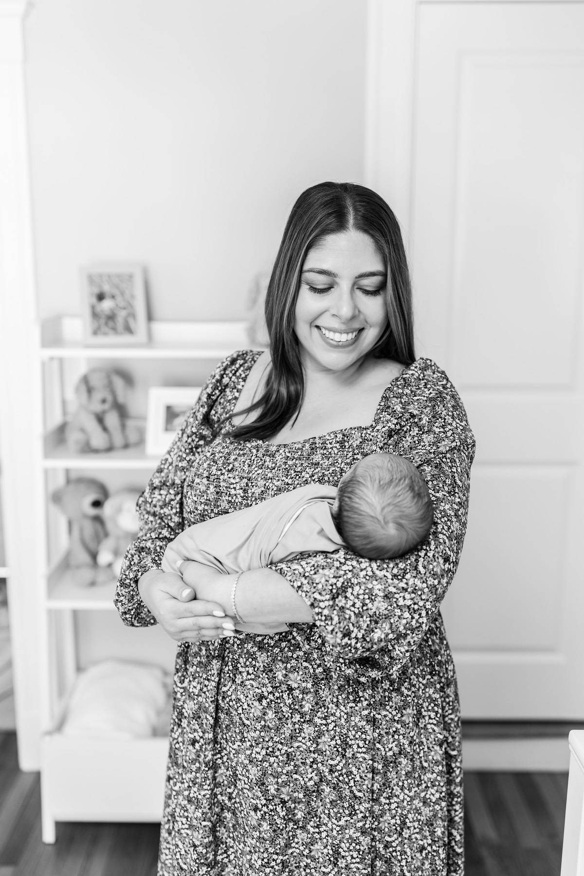 Newborn photo shoot at home of a smiling mother in a floral dress standing in the nursery and cradling her sleeping swaddled newborn baby in black and white