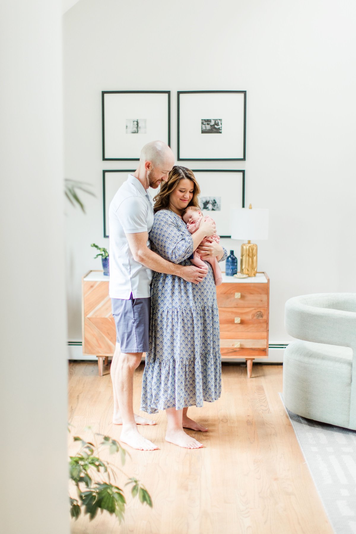 Newborn photos at home of parents standing together in a bright modern living room with gallery wall as mom holds their newborn baby in a pink swaddle and dad wraps his arms around them