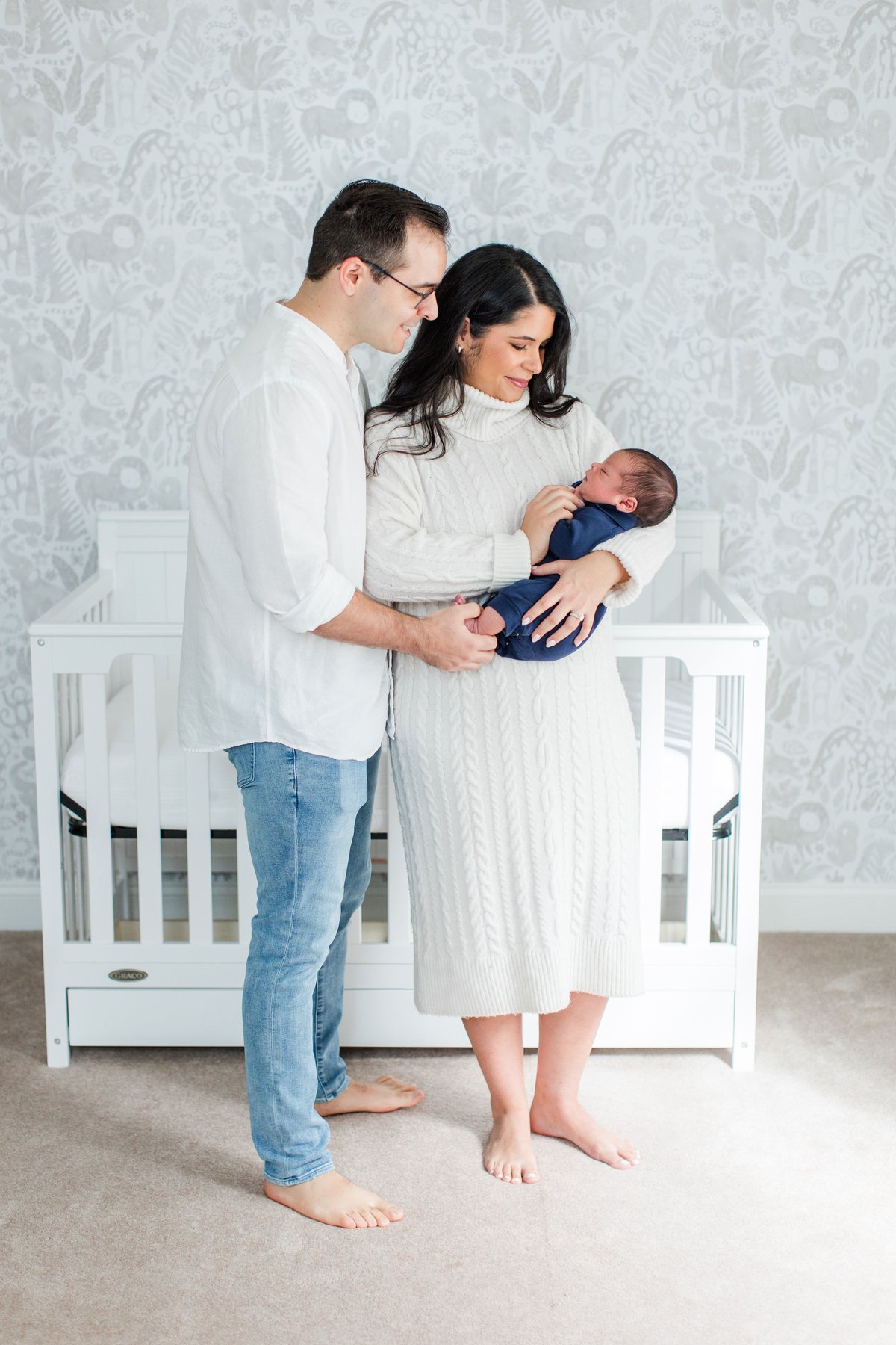 Newborn photos at home of parents standing together in front of a white crib and soft gray animal print wallpaper as mom holds their newborn baby boy in a navy swaddle