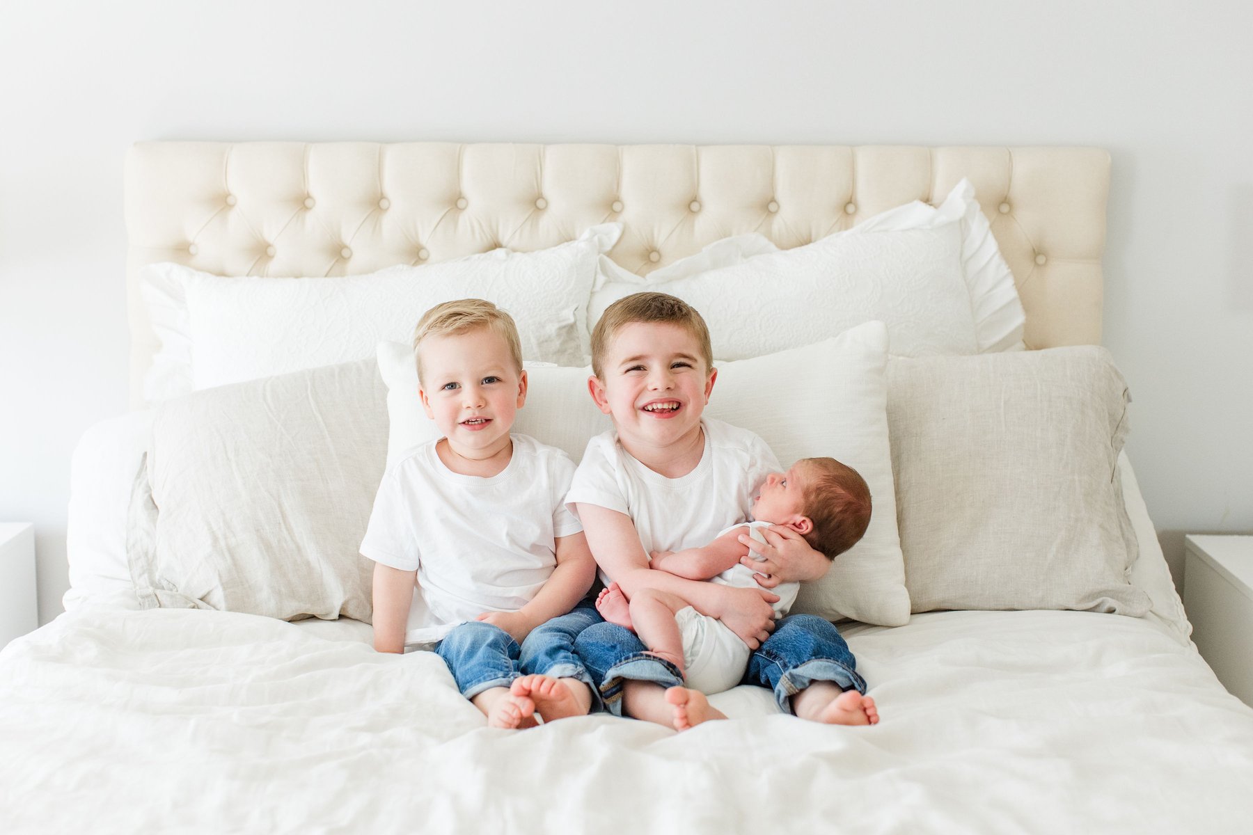 Newborn photos at home of two smiling big brothers in white shirts and jeans sitting on a bed together, with the older brother proudly holding the sleeping newborn baby