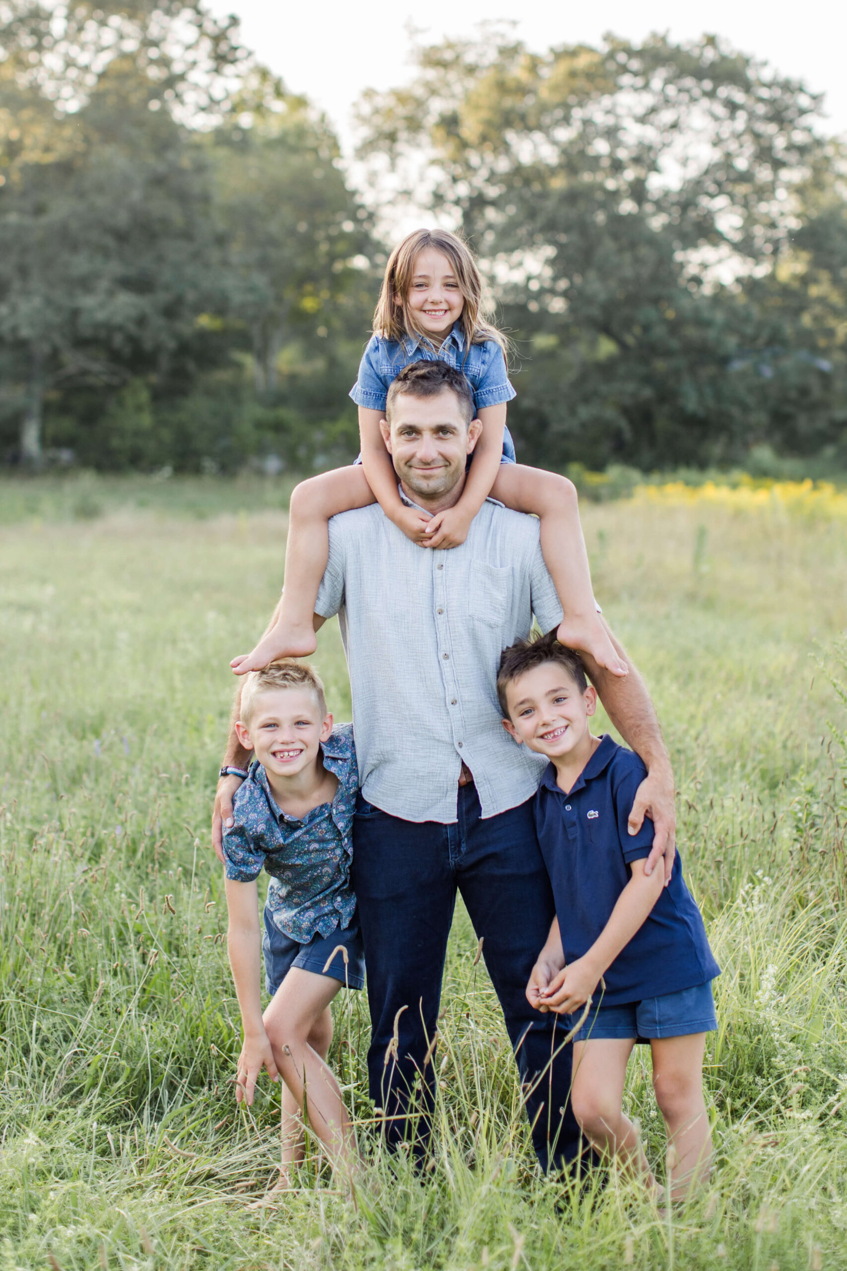 A father poses with his three children in a sunlit meadow during family photos in Boston at golden hour. His daughter sits perched on his shoulders while his two sons lean into him on either side, all four smiling at the camera with wildflowers and glowing green trees stretching behind them.