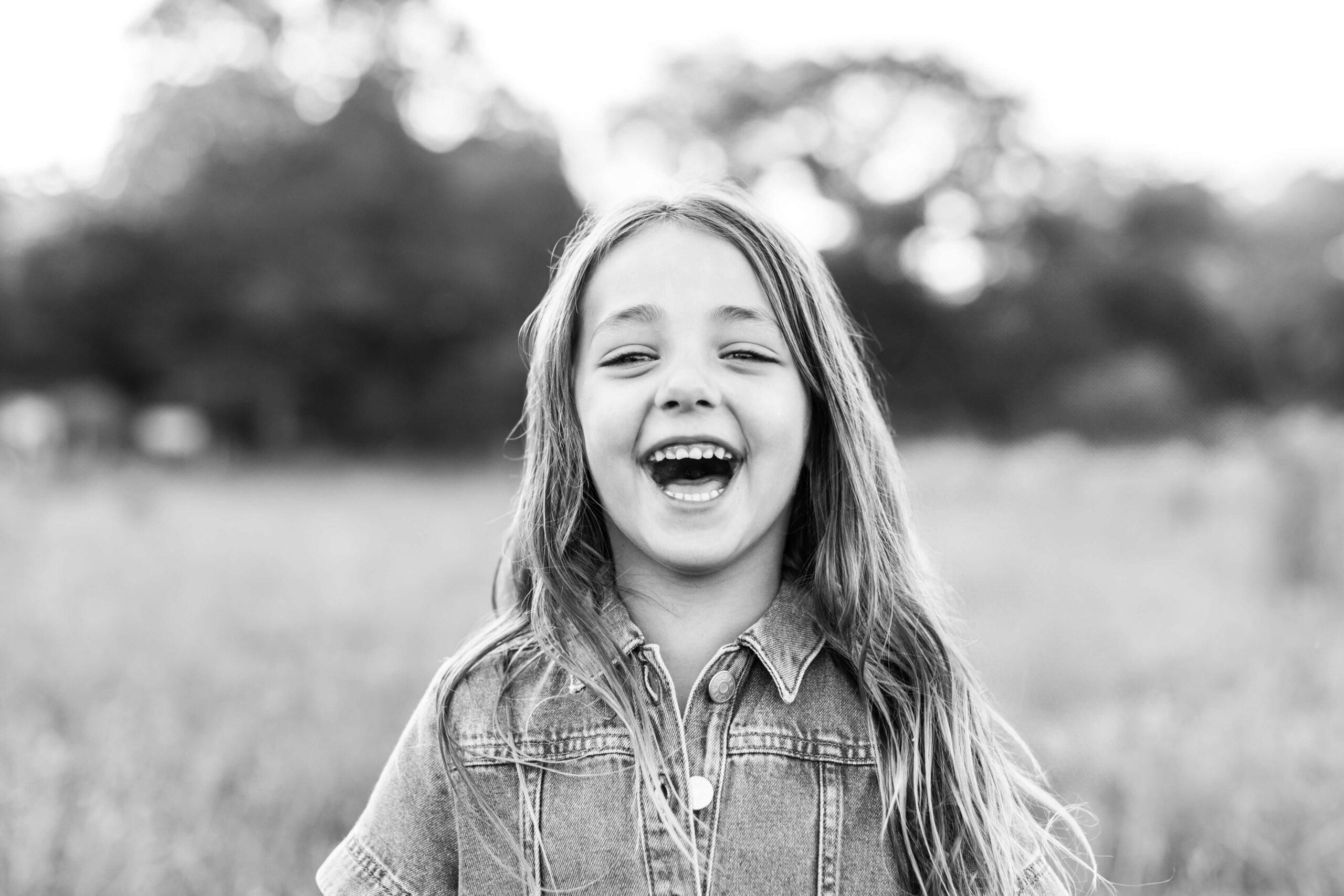 A black and white close-up portrait of a young girl laughing with pure joy during family photos in Boston, her long hair blowing freely. She wears a denim jacket and stands in an open field with softly blurred trees and meadow grass behind her.