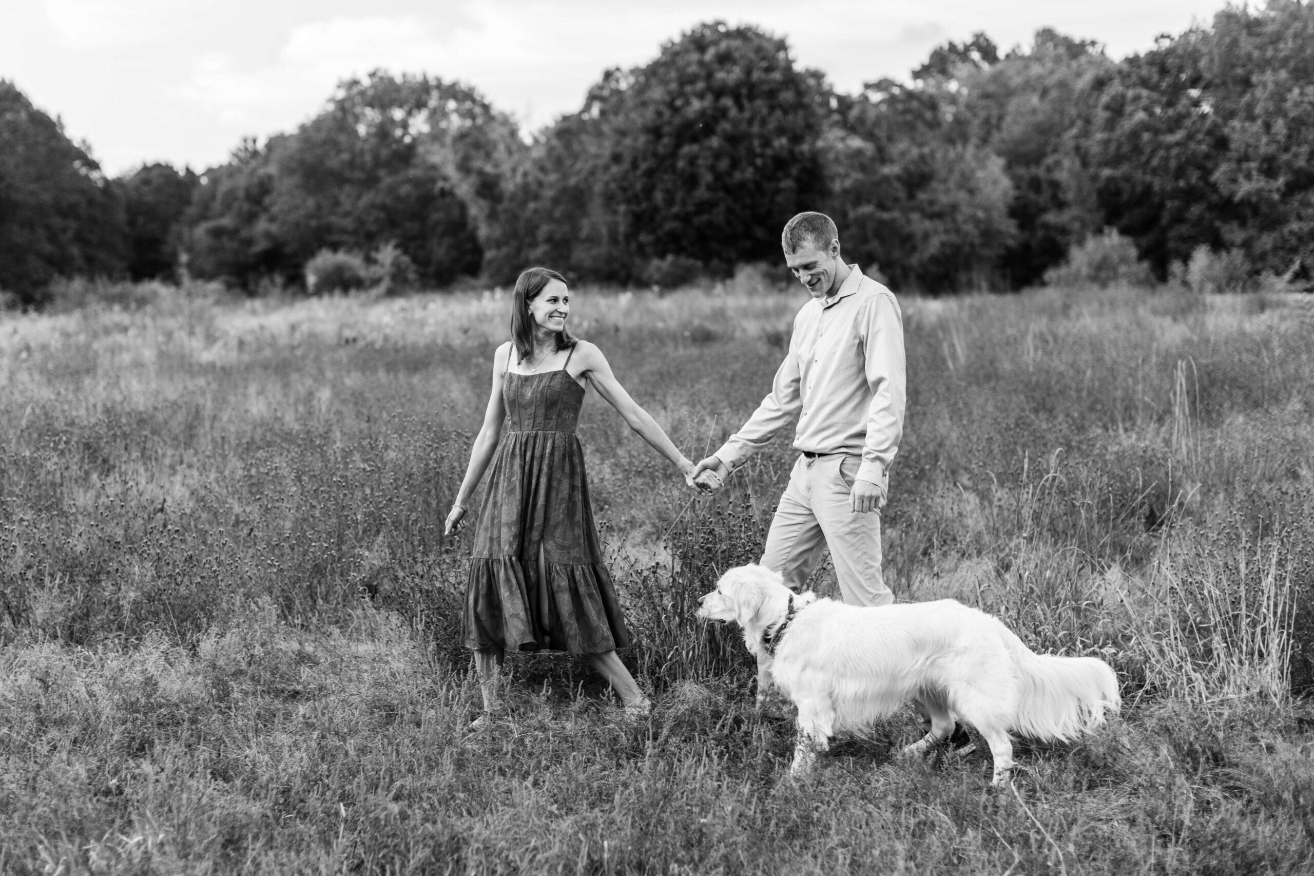 Black-and-white portrait of a couple walking hand in hand through an open field with their golden retriever during a relaxed dog photo shoot.