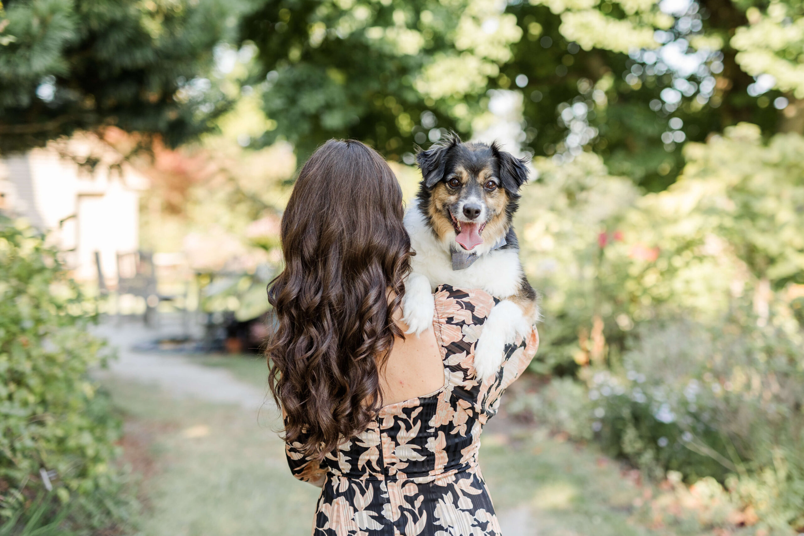 Woman holding a black, white, and tan dog outdoors in a garden with trees and greenery in the background. The dog is looking toward the camera while wearing a gray bow tie.