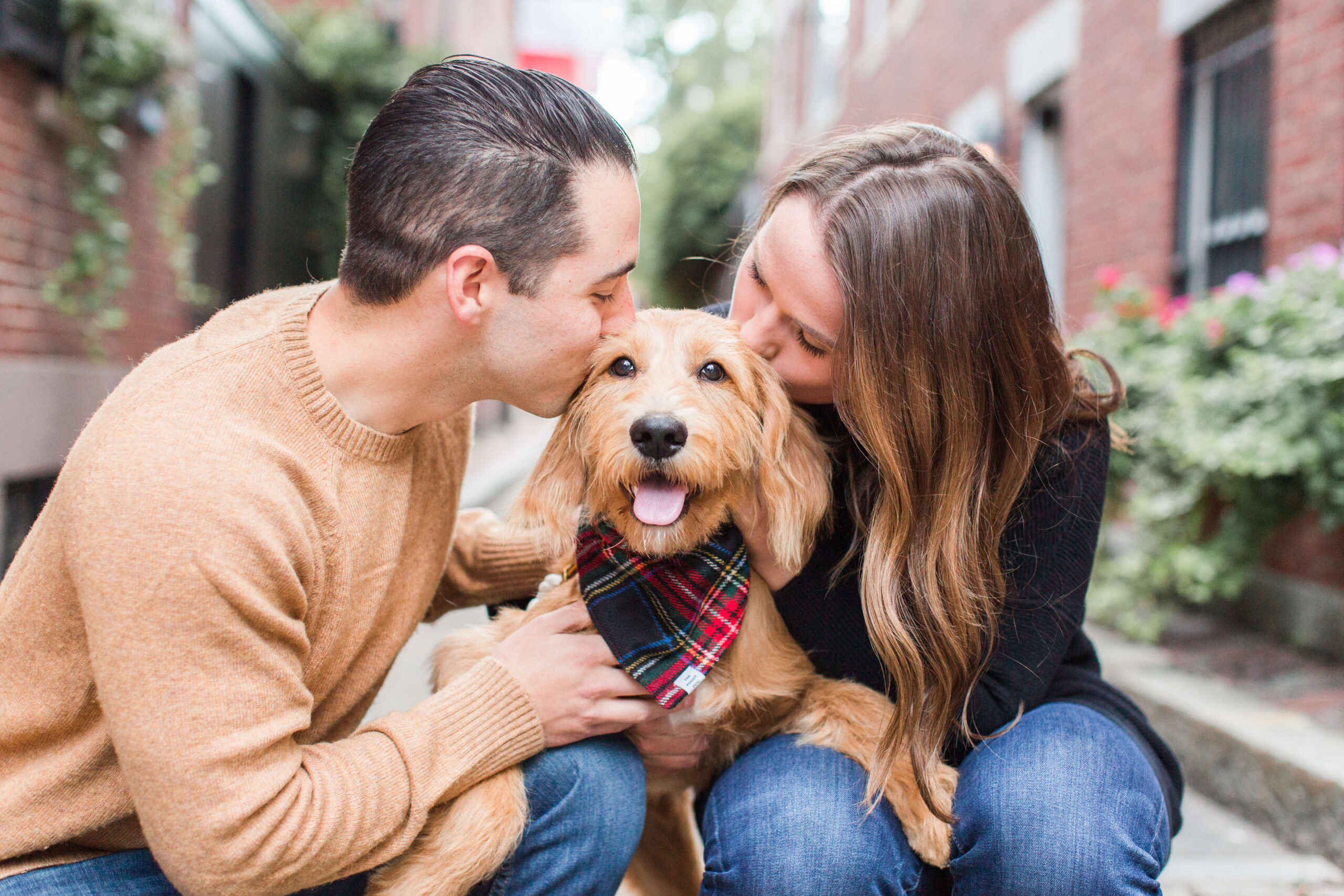 A man and woman kissing a golden doodle wearing a plaid bandana on both cheeks during their family photos