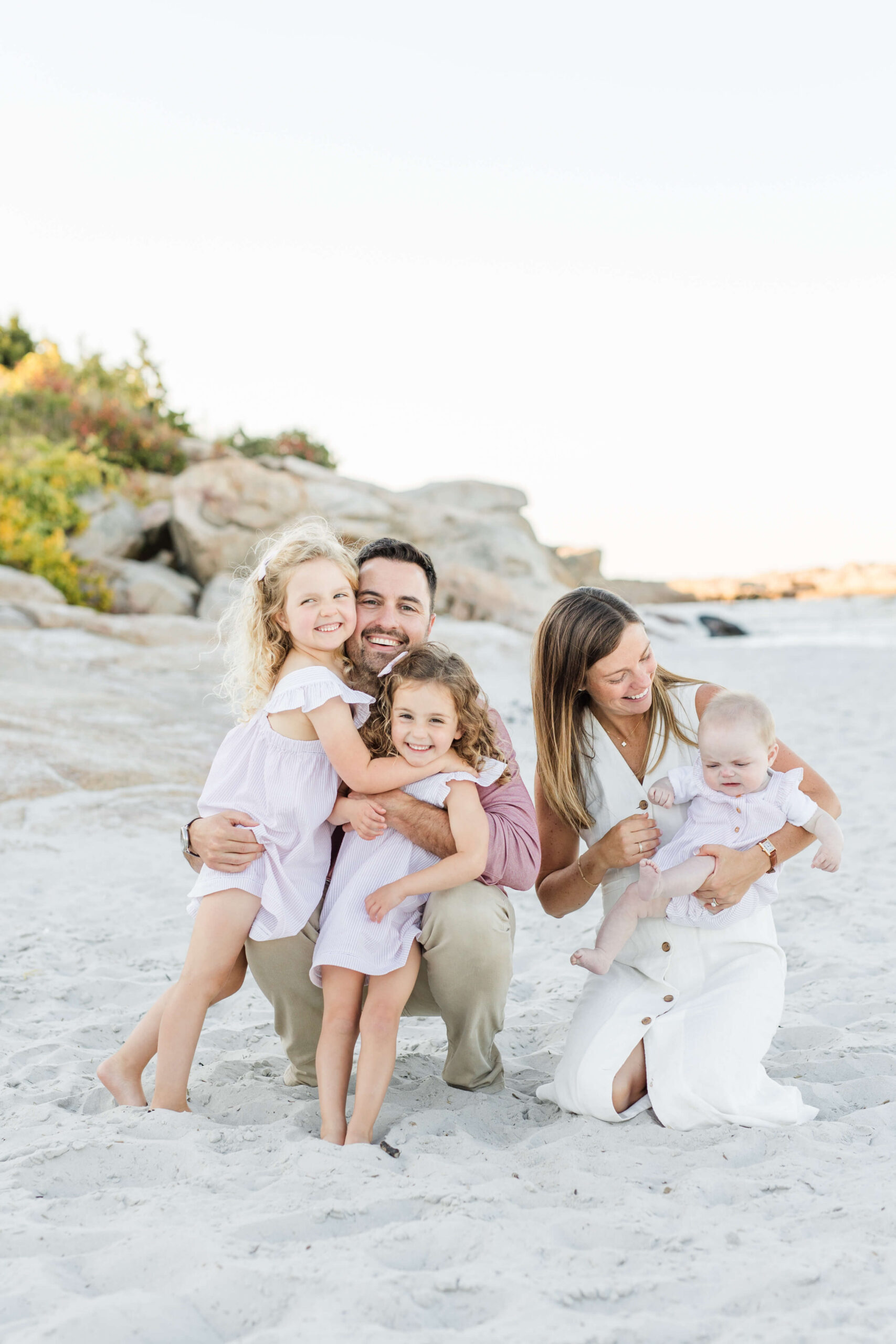 A family of five enjoys a joyful moment together on a sandy beach during a Boston family photo shoot at sunset. The father crouches as his two curly-haired daughters in matching pink dresses hug him, while the mother kneels nearby holding their smiling baby boy, with rocky coastal bluffs in the background.