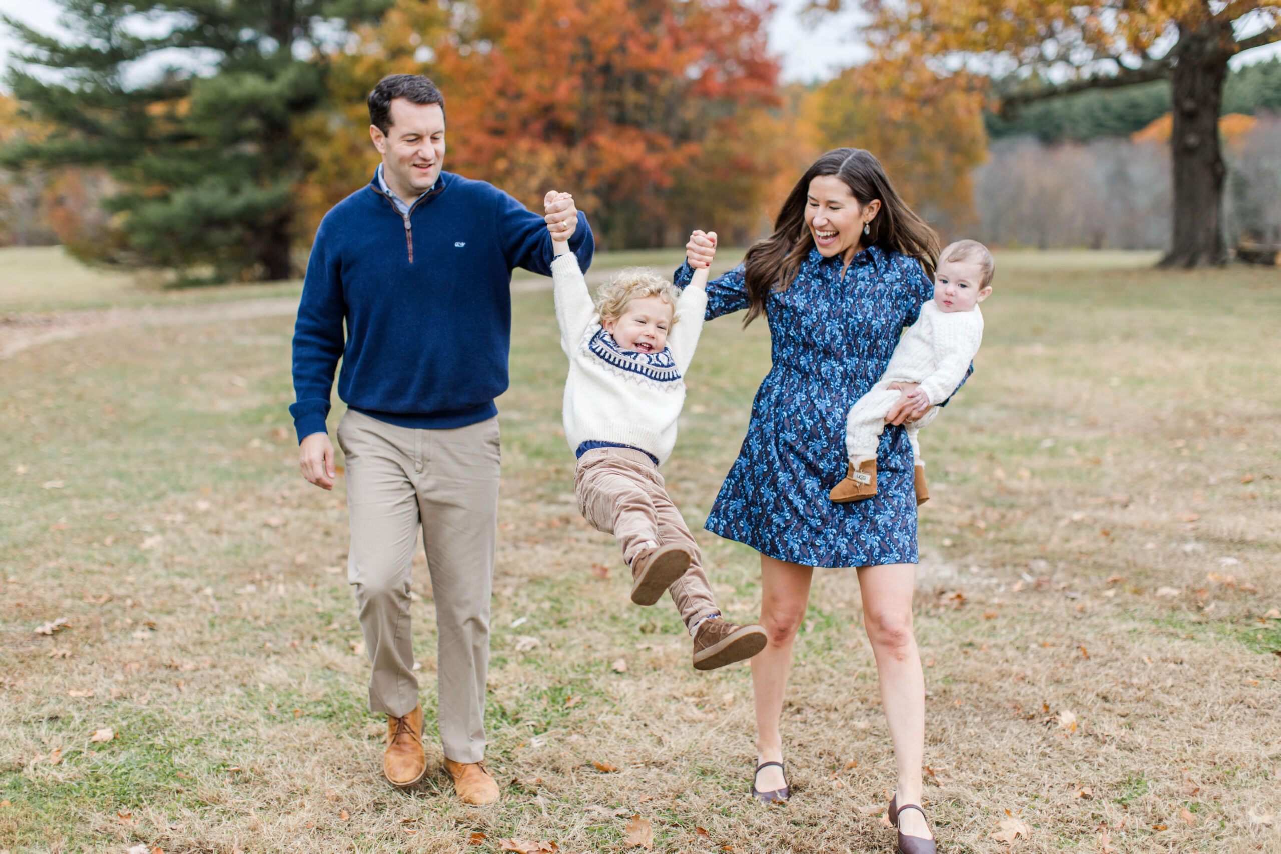 A family of four laughs together during a Boston family photo shoot in a wide-open field bursting with autumn color. The mother holds their baby on one hip while swinging their gleeful curly-haired toddler by the arm, his feet lifting off the ground, as the father walks beside them smiling.