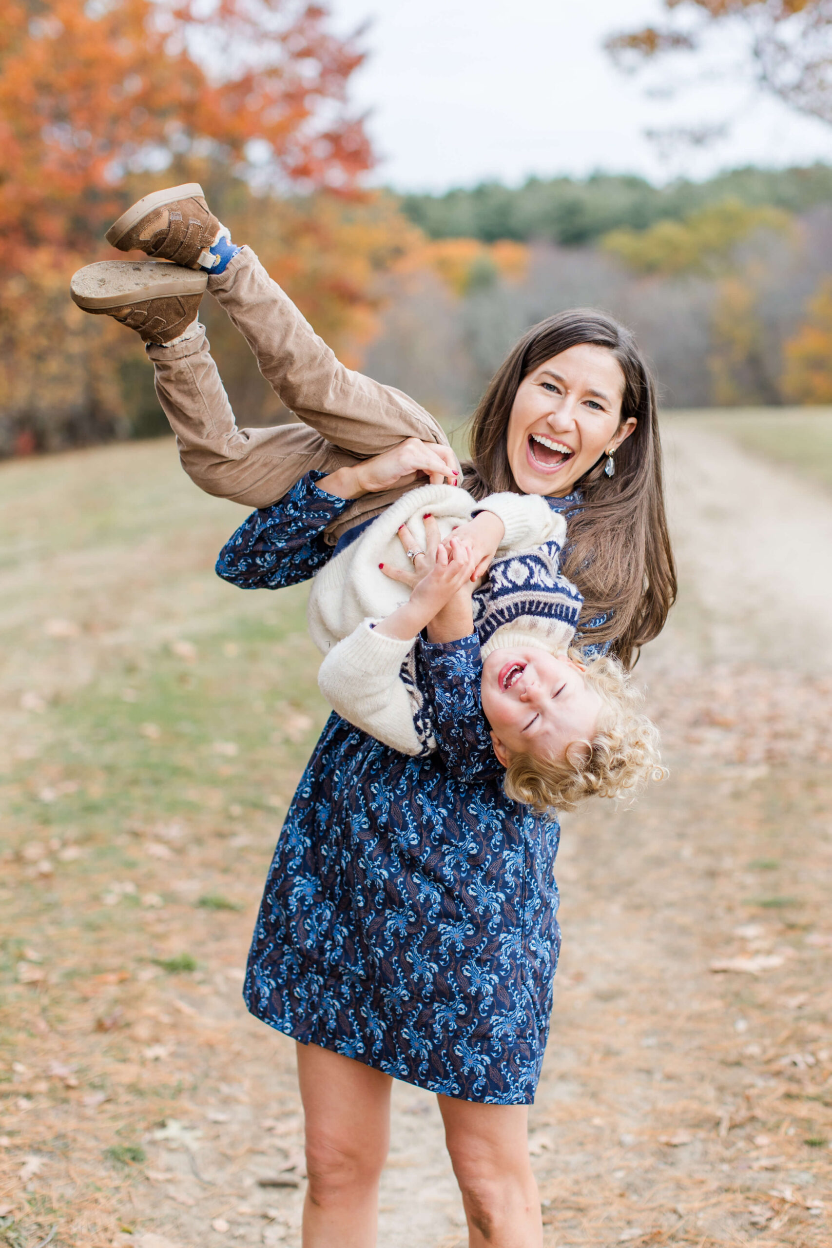 A mother laughs with pure joy as she playfully holds her curly-haired toddler son upside down during a Boston family photo shoot in the fall. Both are laughing as his legs fly in the air, with a dirt path and colorful autumn trees stretching behind them.