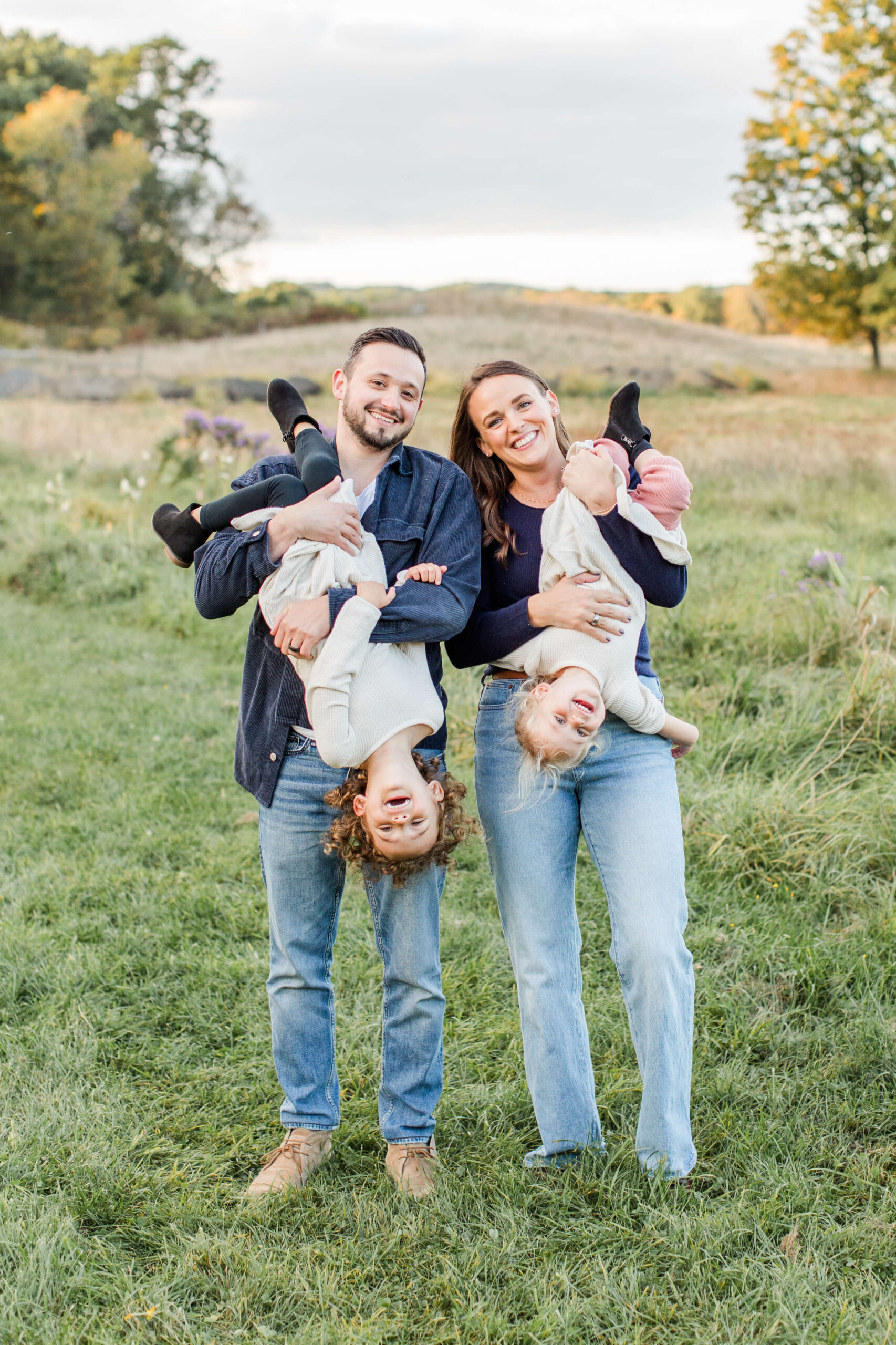 Two parents each hold one of their young children upside down during a Boston family photoshoot in an open field, all four laughing together. Both kids hang gleefully with their legs in the air as their parents smile at the camera, with rolling hills, wildflowers, and early autumn trees in the background.