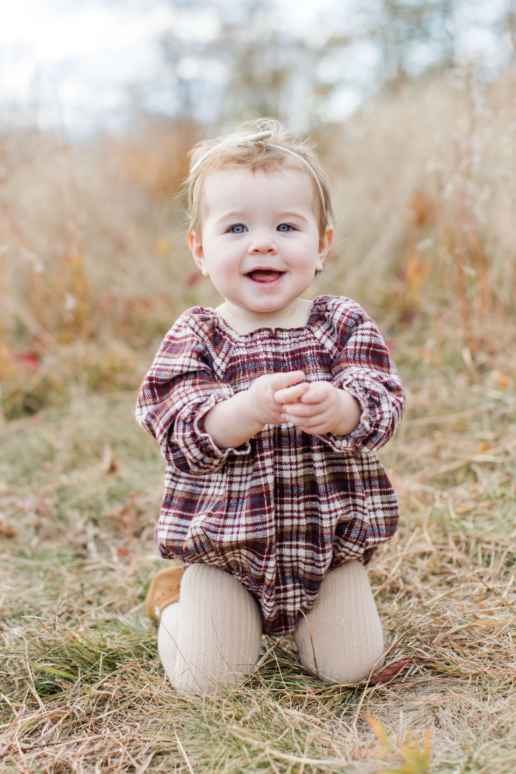 A blue-eyed baby girl kneels in a golden autumn meadow during Boston kid portraits, clapping her hands together with a big grin. She wears a cozy burgundy plaid bubble romper with cream tights, with dried grasses and a soft blurred sky behind her.