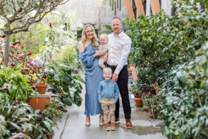 A family of four poses together during a family photo shoot inside a lush botanical conservatory, surrounded by tropical plants and terracotta pots. The mother in a blue satin dress holds their smiling baby while their young blond son stands in front with the father, all dressed in coordinating shades of blue.