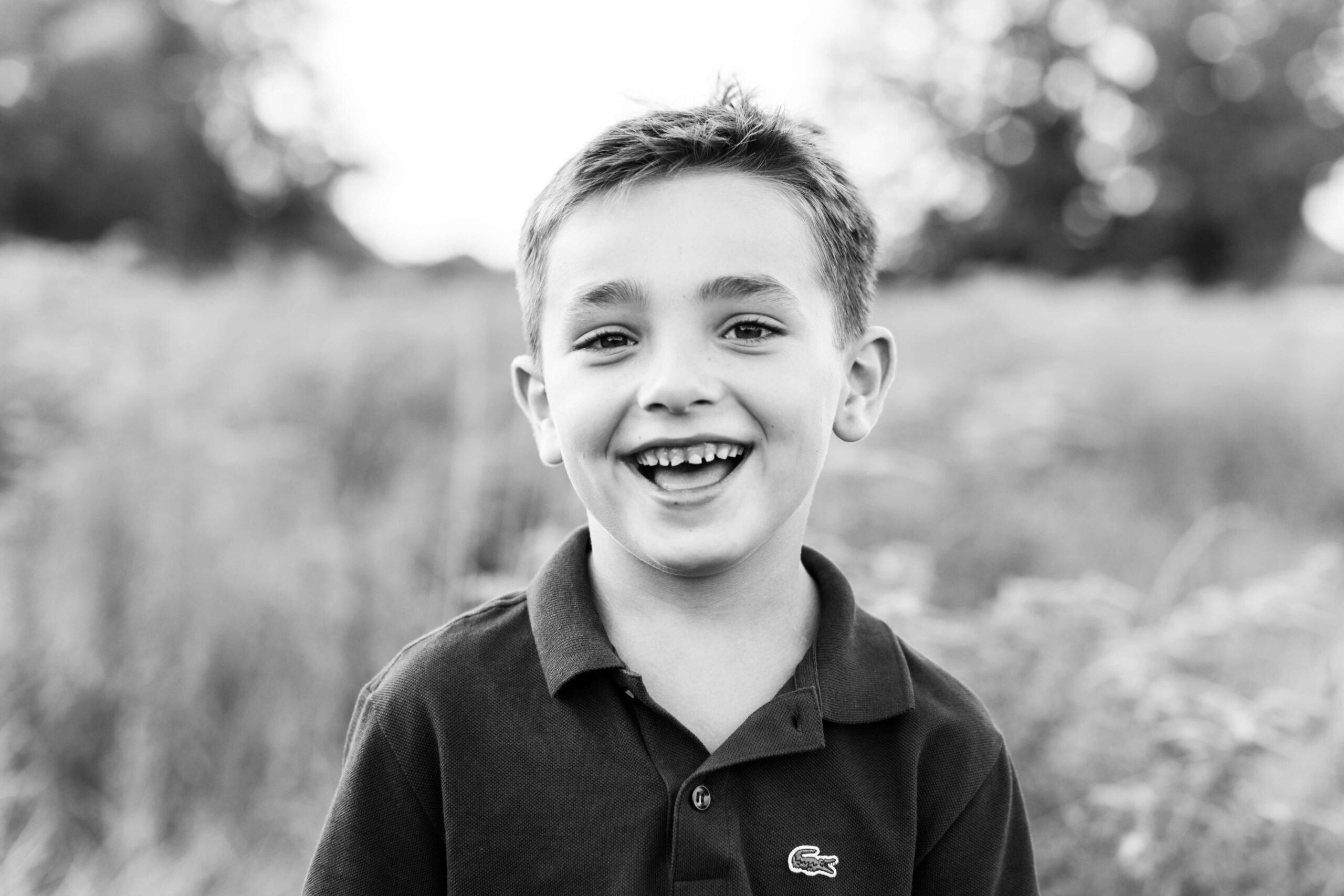 A black and white close-up portrait of a young boy laughing with a wide smile during a family photo shoot in Boston, wearing a Lacoste polo shirt. Tall meadow grasses and bokeh light create a soft, dreamy background behind him.