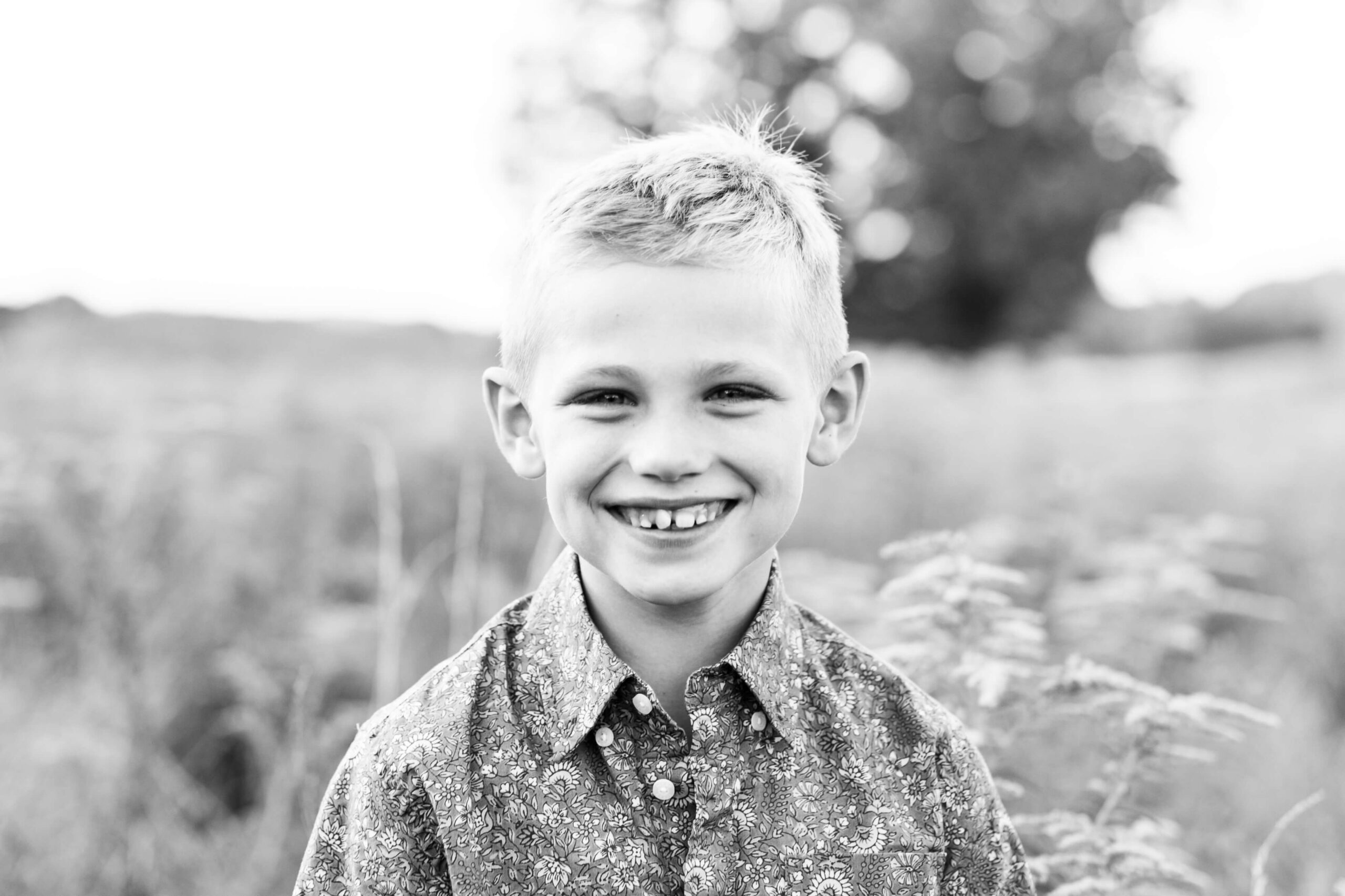 A black and white close-up portrait of a young boy grinning broadly during a family photo shoot in Boston, wearing a floral button-down shirt. He stands in an open field with tall grasses and a large tree softly blurred in the background.