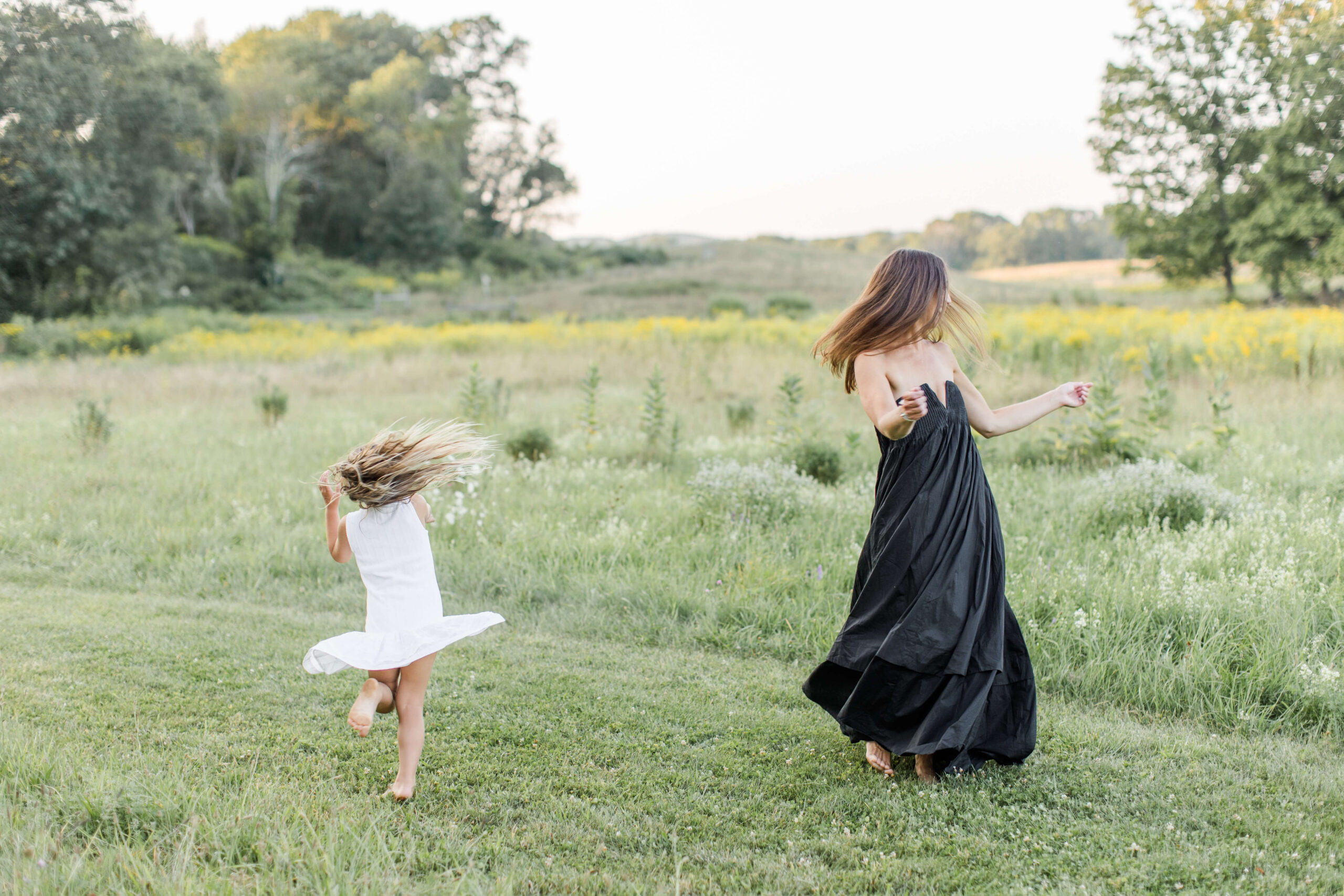 A mother and young daughter twirl and dance barefoot in an open wildflower meadow during a family photo shoot, their hair and dresses caught mid-spin. Seen from behind, the mother wears a flowing black gown while her daughter wears a white dress, with yellow wildflowers and trees glowing in the warm evening light.