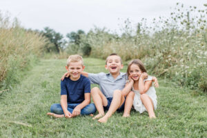 Three siblings sit barefoot on a grassy path during family photos in Boston, laughing and smiling at the camera. The middle boy drapes his arms around his blonde brother on the left and his younger sister on the right, with tall wildflowers and meadow grasses framing them on either side.