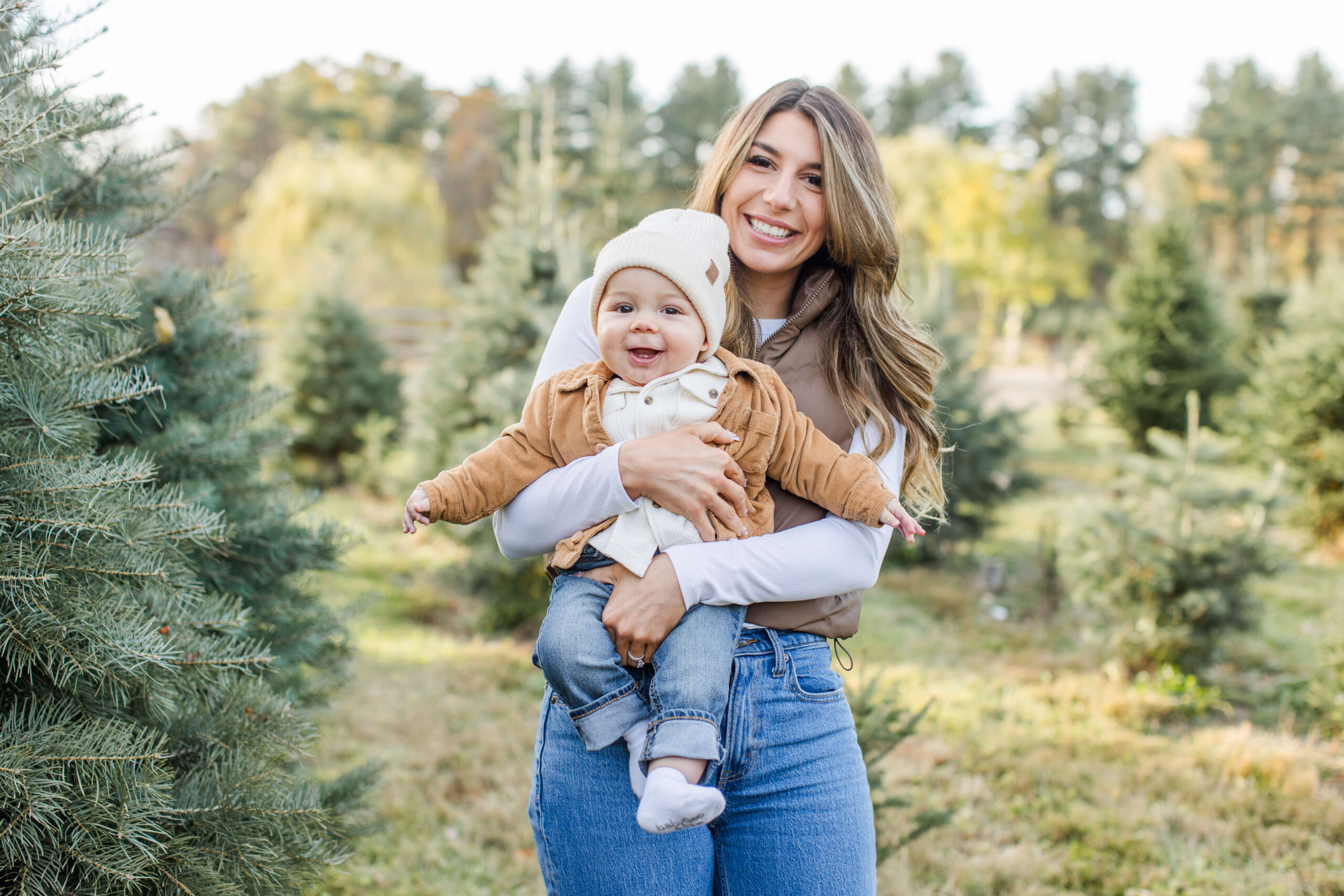A mother holds her smiling baby boy during a family photoshoot at a Christmas tree farm, both looking at the camera with big grins. The baby wears a cream knit beanie and camel corduroy jacket as his mother holds him against a backdrop of lush green pine trees and soft autumn foliage.