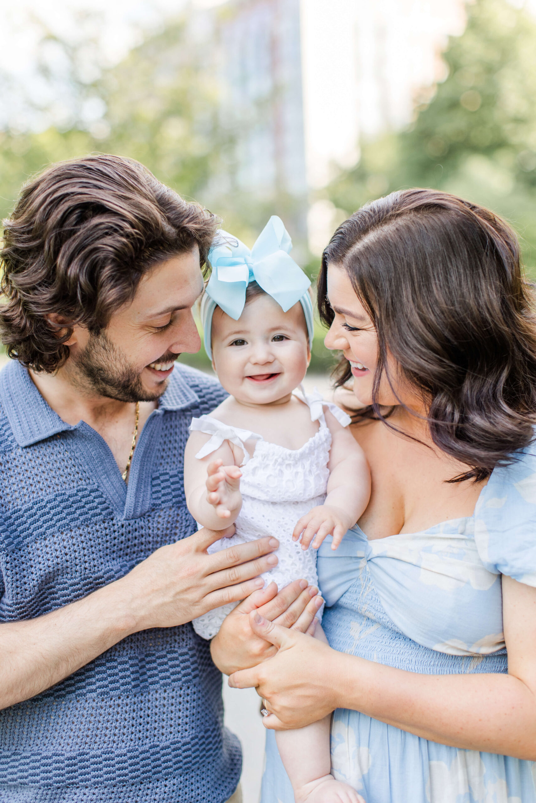 A close-up portrait of two parents smiling down at their baby girl during a photoshoot with kids, all dressed in coordinating shades of blue. The beaming baby wears a large light blue bow headband and a white eyelet dress as she looks straight at the camera, held between her parents against a soft green background.