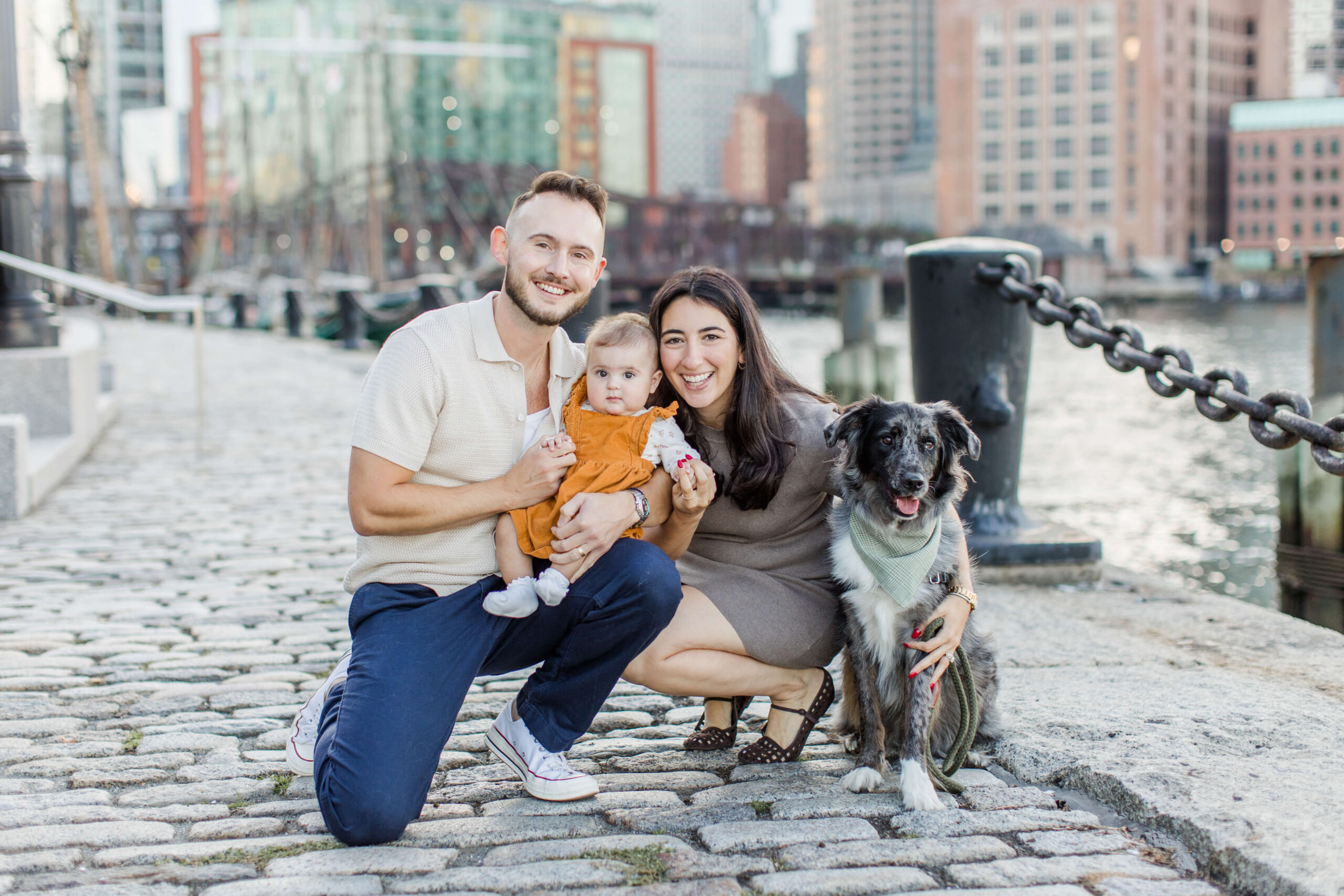 Family posing with their baby and black dog during a Seaport Boston dog photo shoot along the harbor. The family kneels on a cobblestone walkway with city buildings and waterfront views in the background.