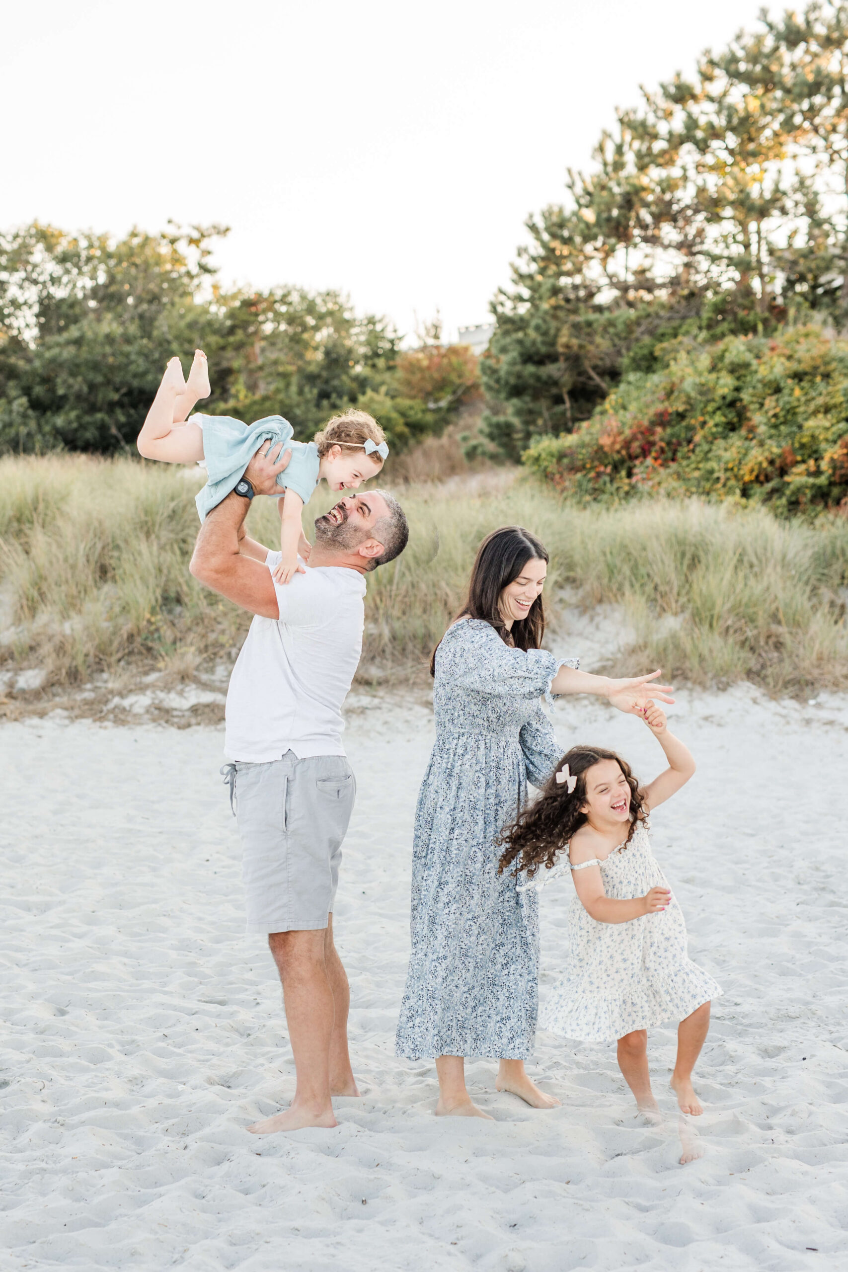 A family of four plays together on a sandy beach during a spring family photo shoot in Boston. The father lifts his giggling toddler daughter high in the air while the mother spins their older curly-haired daughter by the hands, with coastal dune grasses and autumn-tinged shrubs in the background.