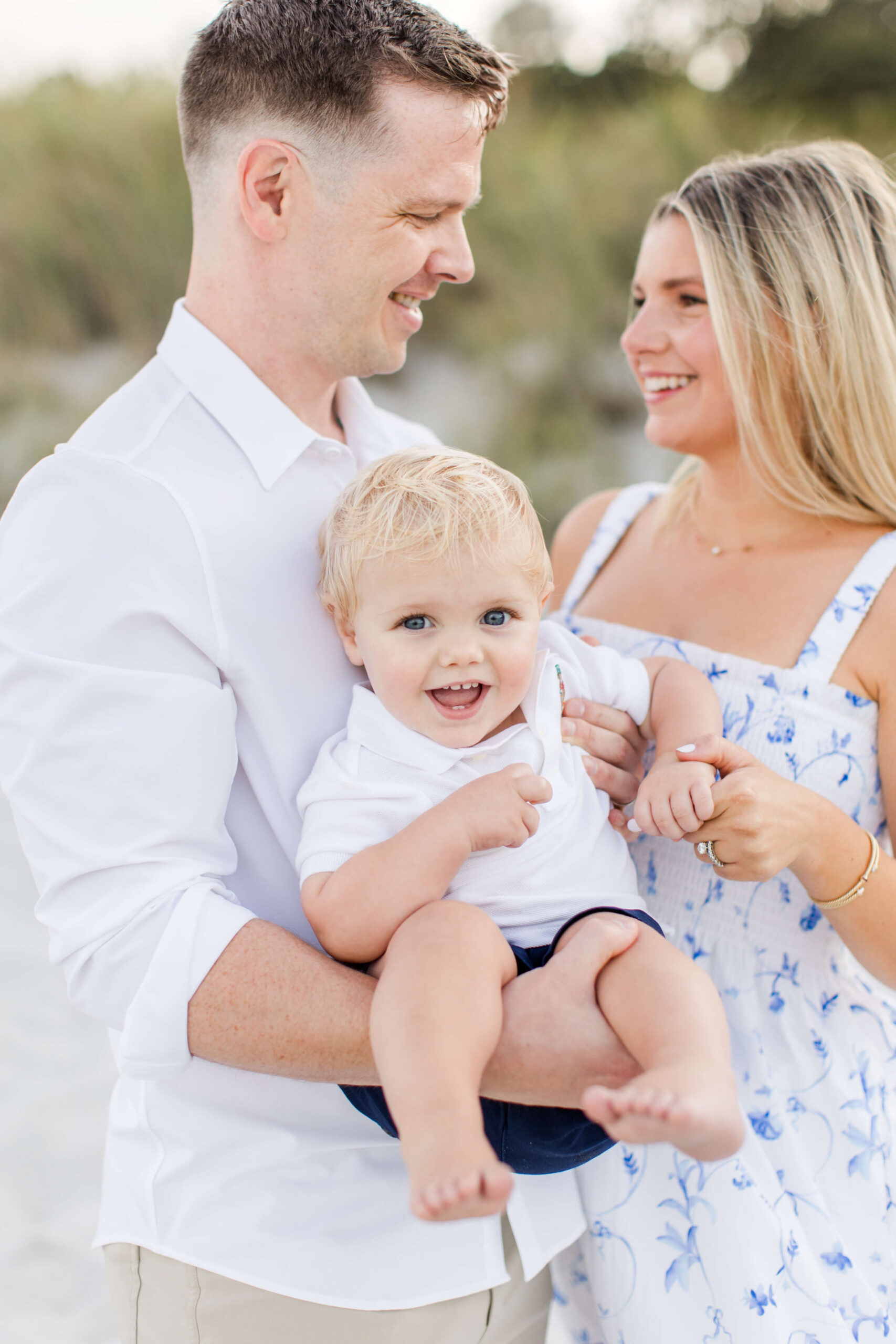 A family of three poses together on a park path during a photoshoot with kids in an urban garden setting. The mother in a floral light blue maxi dress holds their baby girl in a matching blue bow headband, while the father in a blue knit polo smiles beside them, with manicured hedges, roses, and city buildings visible in the background.