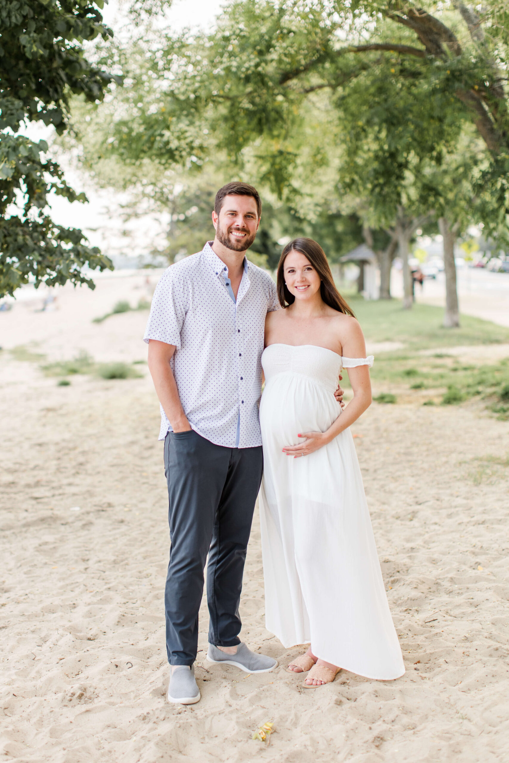 A smiling couple standing together on a sandy beach under trees, the woman in a white off-the-shoulder maxi dress cradling her baby bump