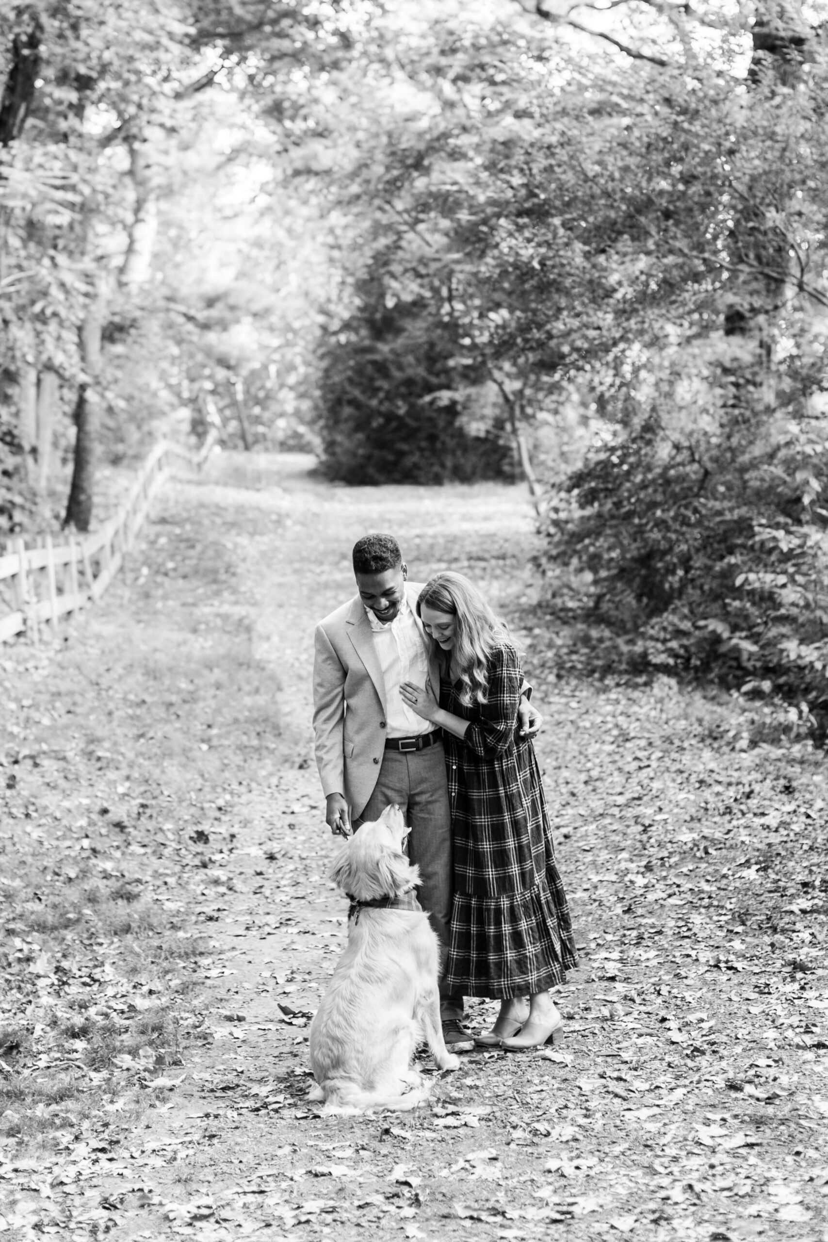 Black-and-white portrait of a couple standing on a tree-lined path with their golden retriever at Arnold Arboretum. The dog looks up at them while fallen leaves cover the ground beneath the canopy of trees.