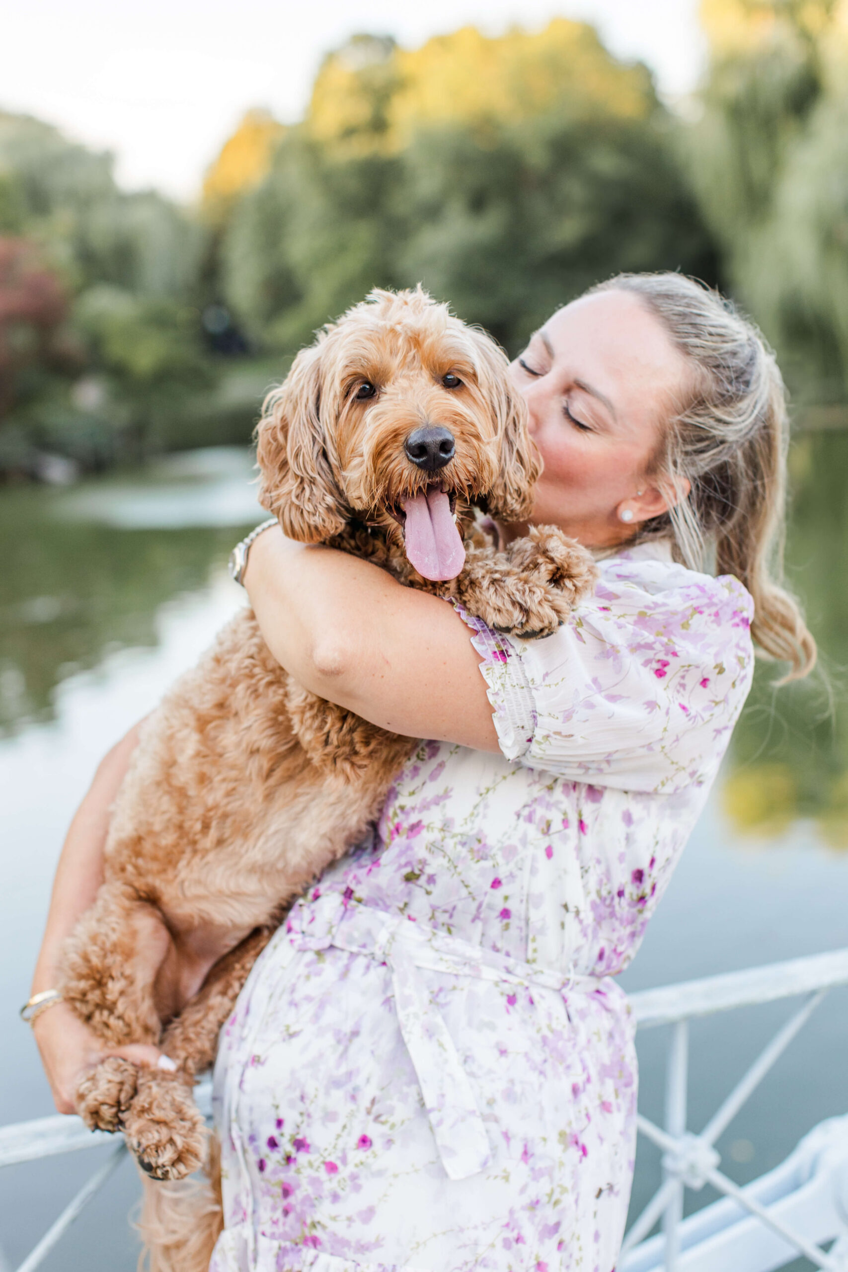 Woman holding and cuddling her apricot-colored doodle during a dog photo shoot at Boston Public Garden. The dog looks toward the camera while the pond and soft green landscape create a natural backdrop.