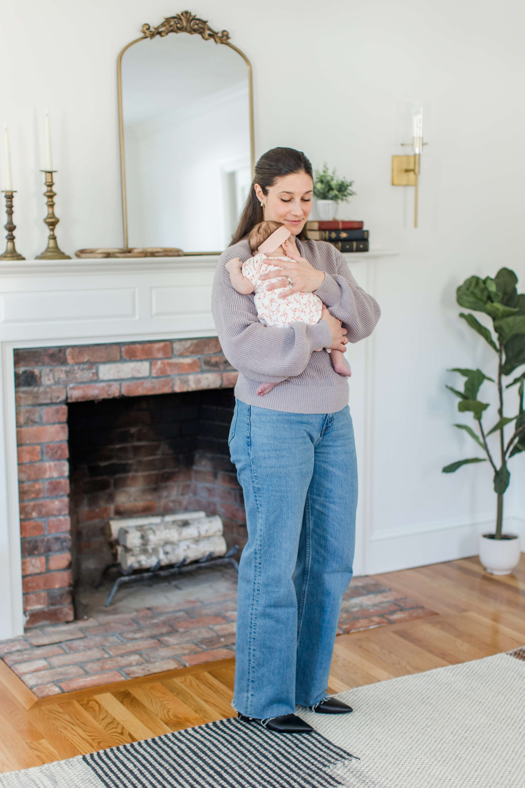 A mother in a lavender sweater and wide-leg jeans standing in front of a brick fireplace with a gold mirror, cradling her newborn baby