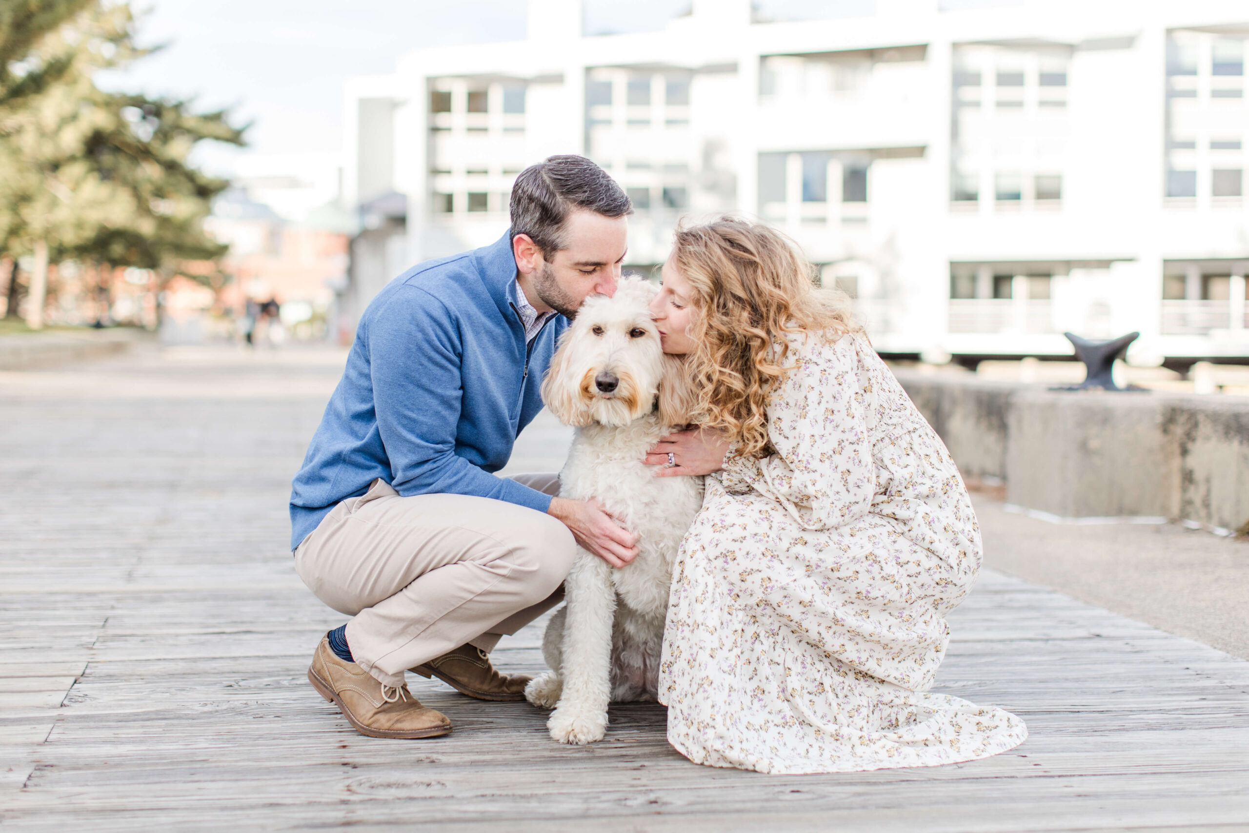 Couple kneeling beside their cream-colored doodle during a dog photo shoot at Charlestown Navy Yard. They cuddle their dog on the wooden boardwalk with waterfront buildings and harbor views in the background.
