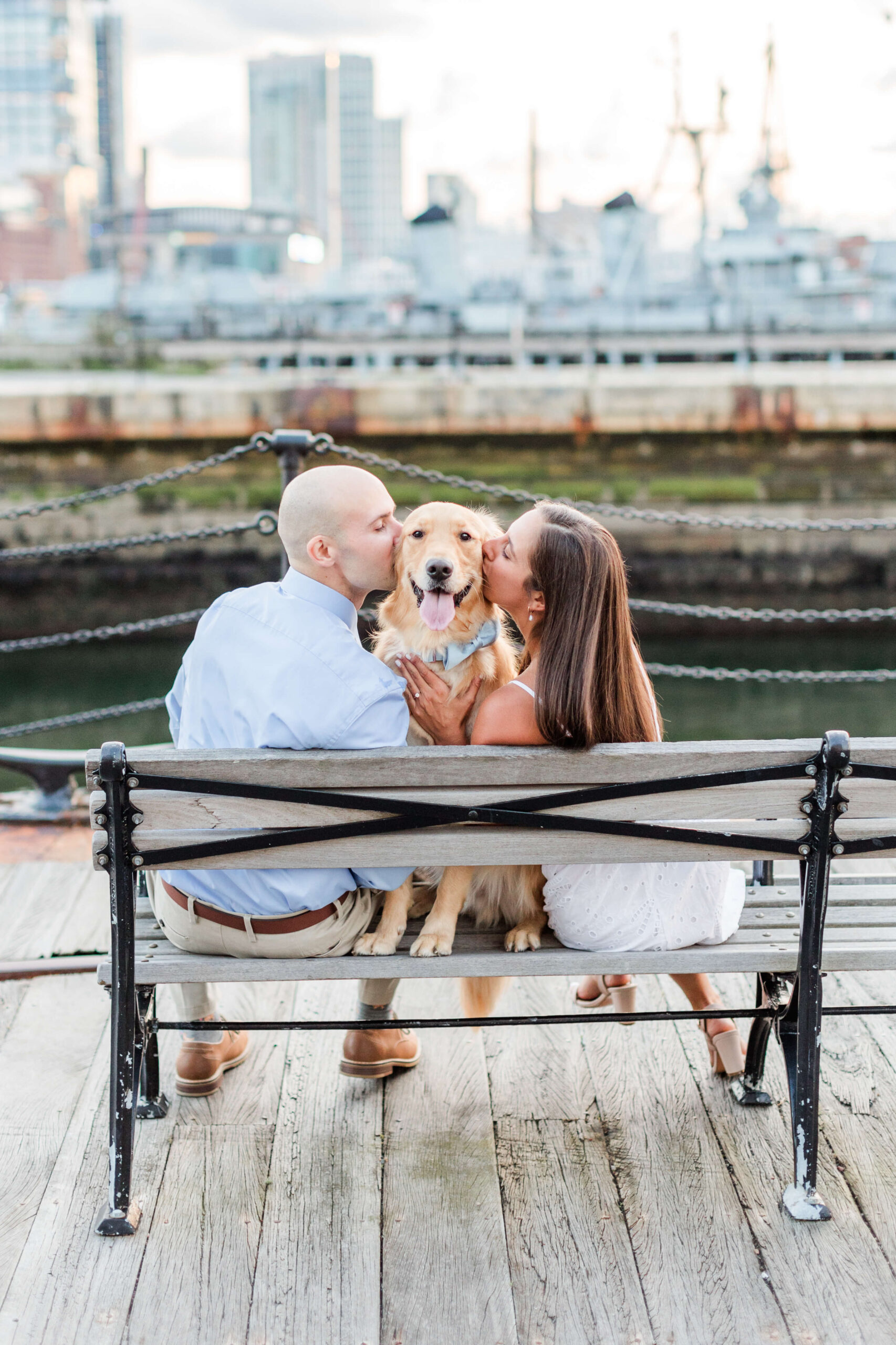 Couple sitting on a waterfront bench with their golden retriever during a dog photo shoot at Charlestown Navy Yard. They kiss the dog on each cheek while harbor views, historic ships, and the Boston waterfront create a scenic backdrop.