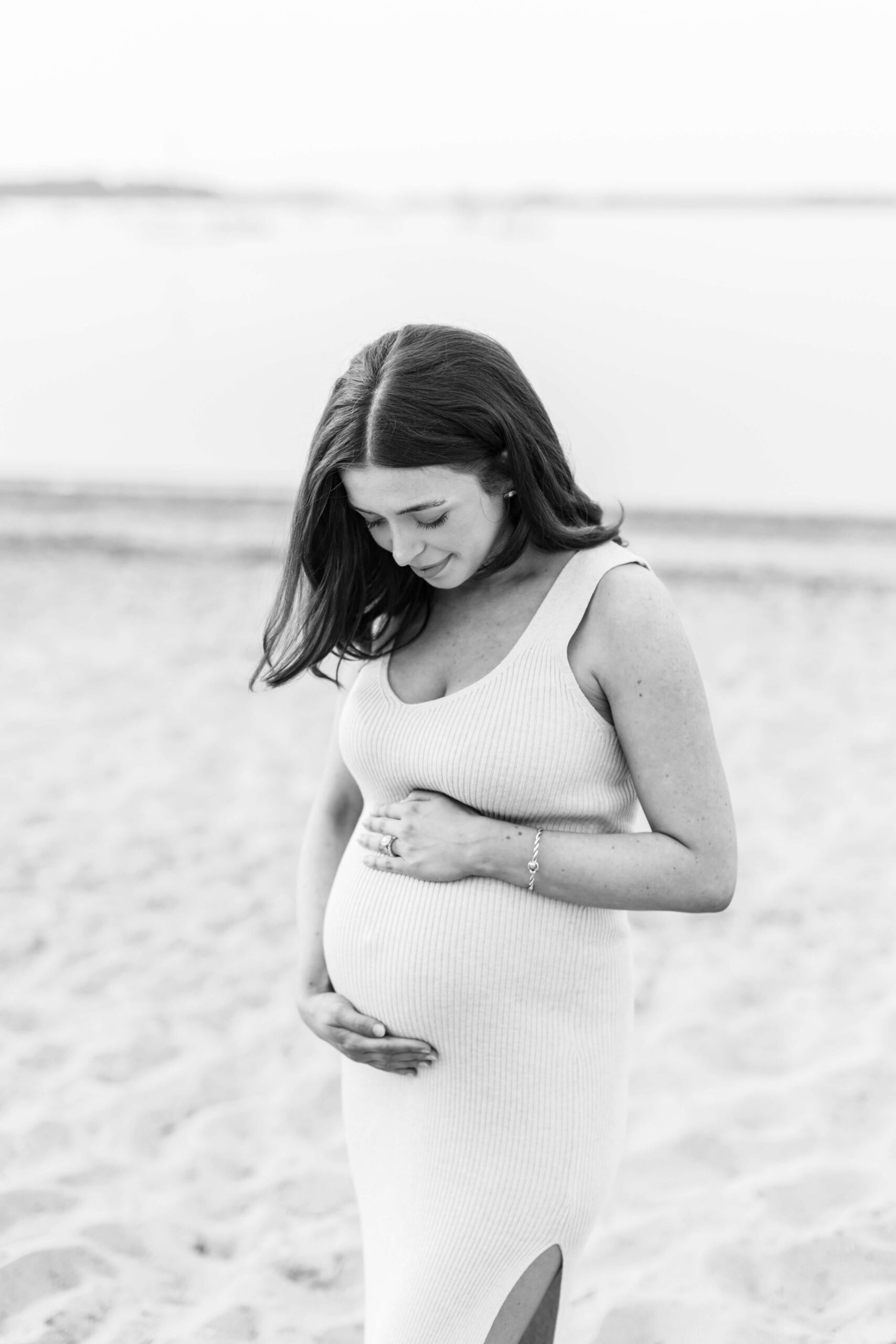 A black and white photo of a pregnant woman in a fitted ribbed dress standing on the beach, looking down and gently holding her bare baby bump