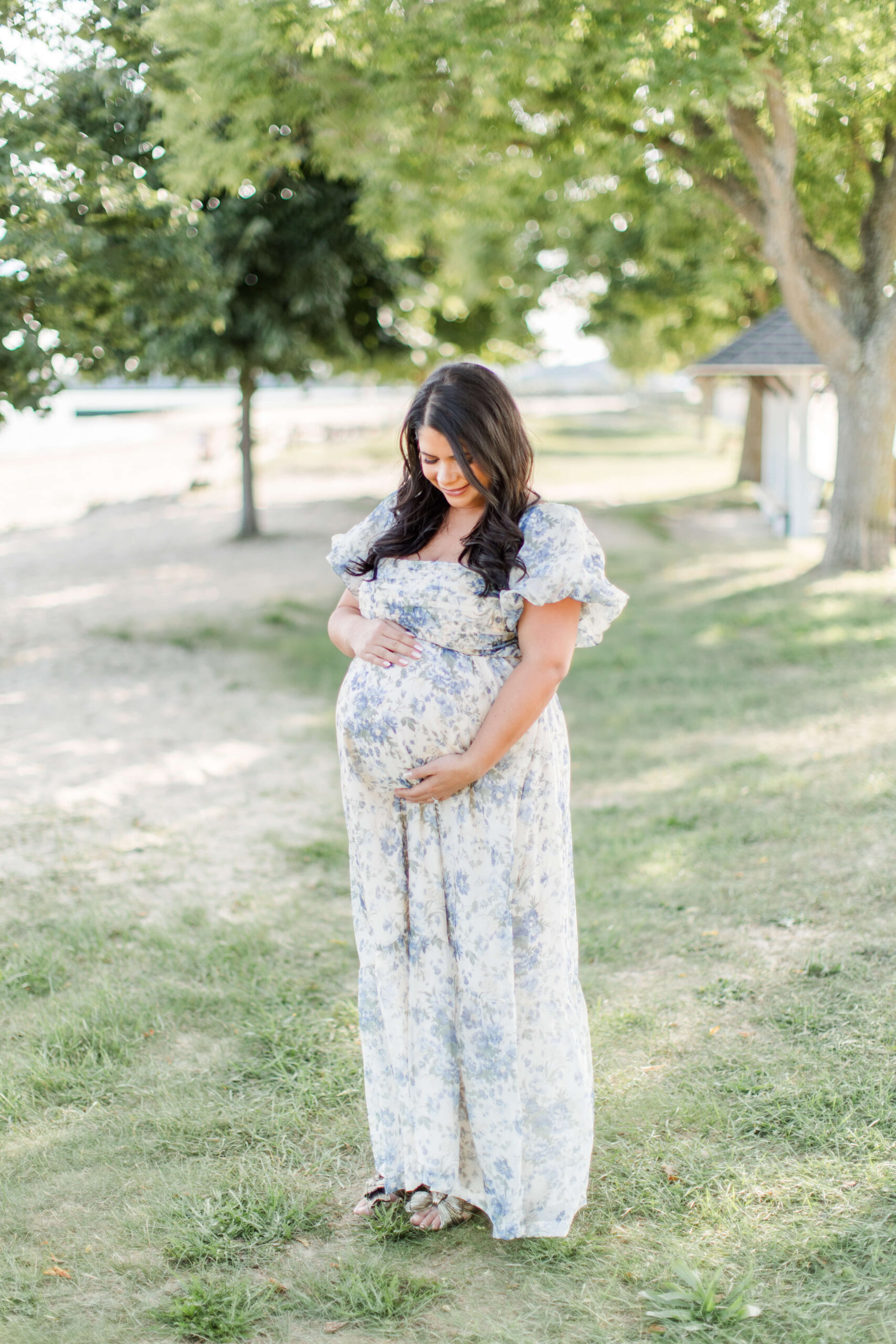 A pregnant woman in a flowing floral dress standing alone outdoors among trees, smiling softly while holding her baby bump