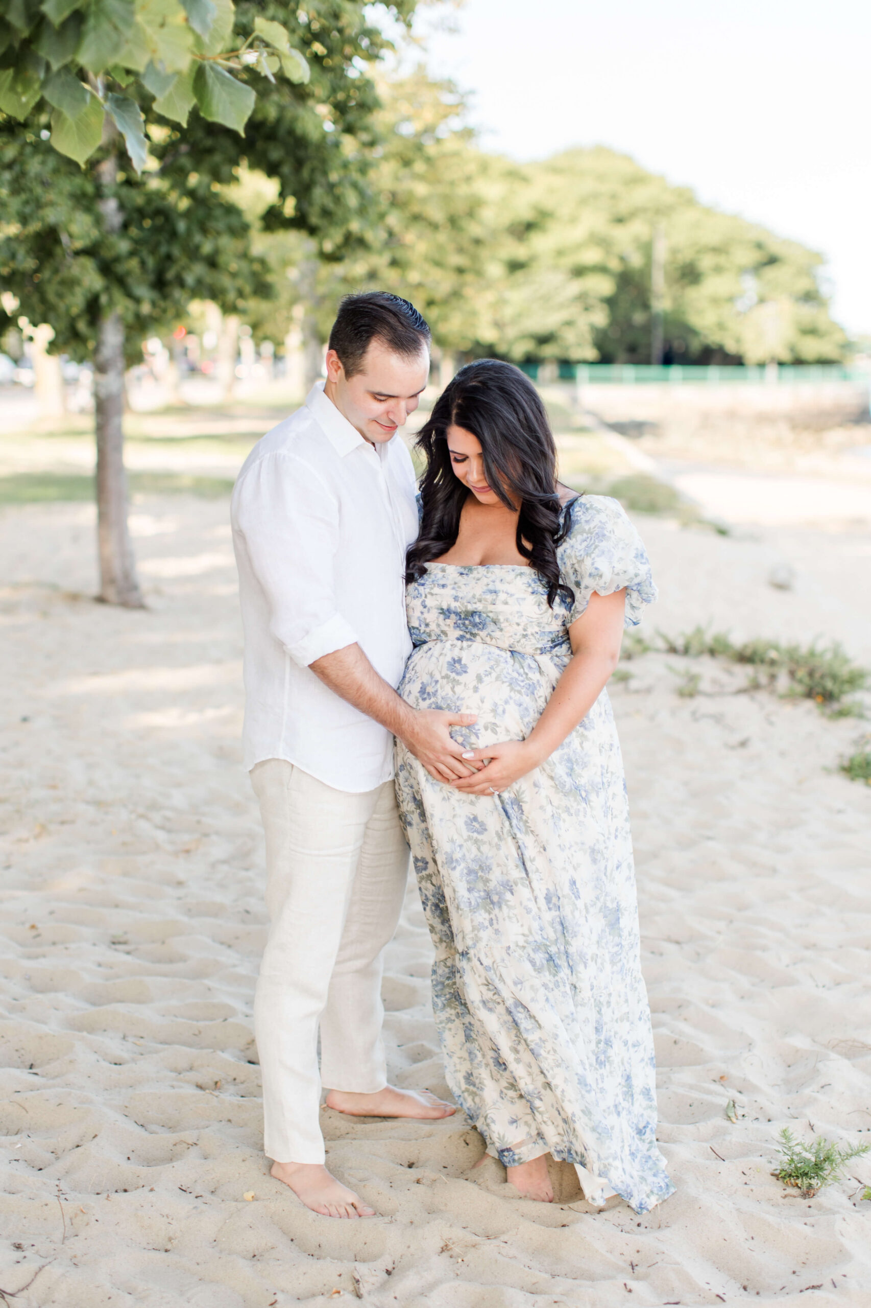 A couple outdoors in soft natural light, the man in a white shirt standing behind the woman in a floral dress as they both look down and cradle her baby bump together