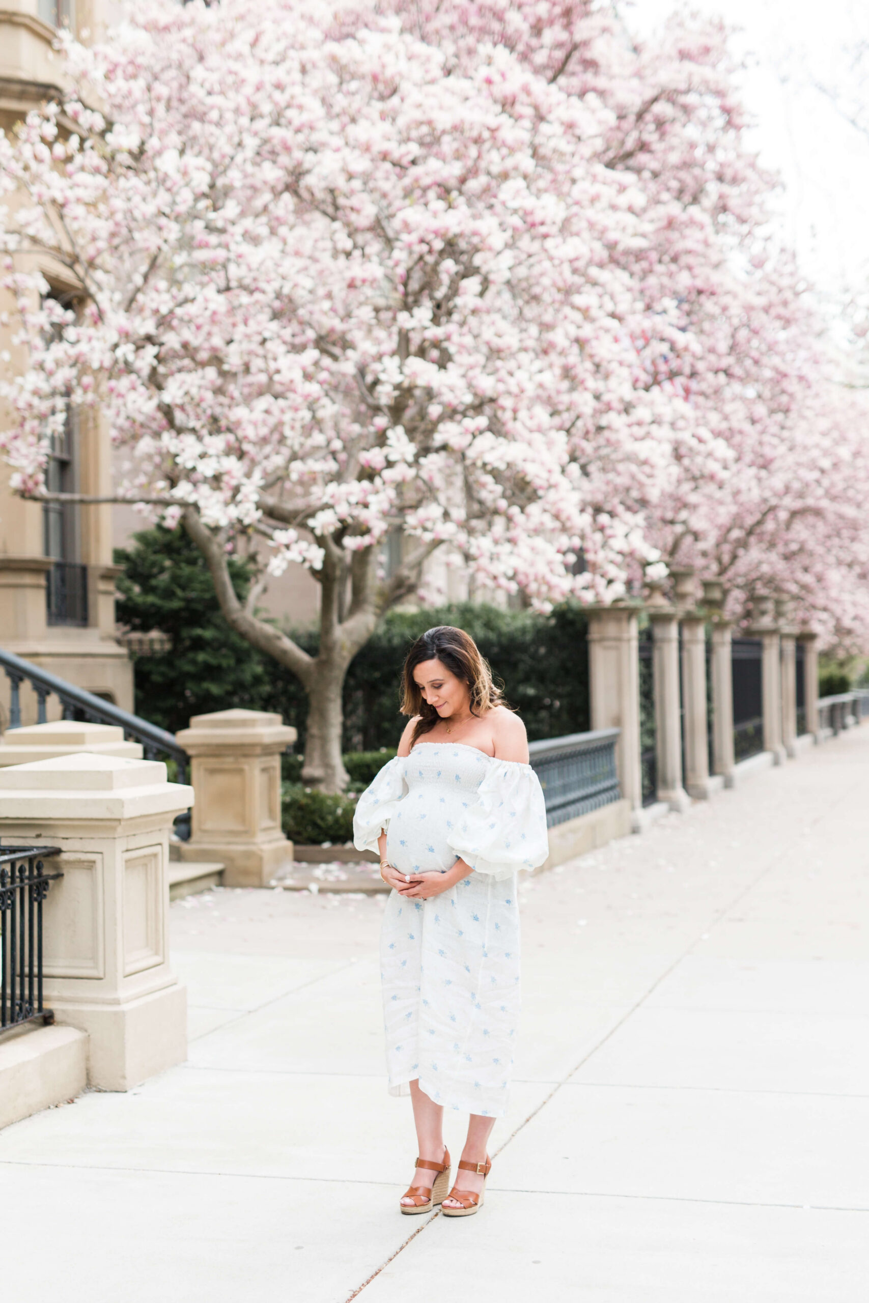 A pregnant woman in a white off-the-shoulder dress standing on a city sidewalk lined with blooming pink cherry blossom trees