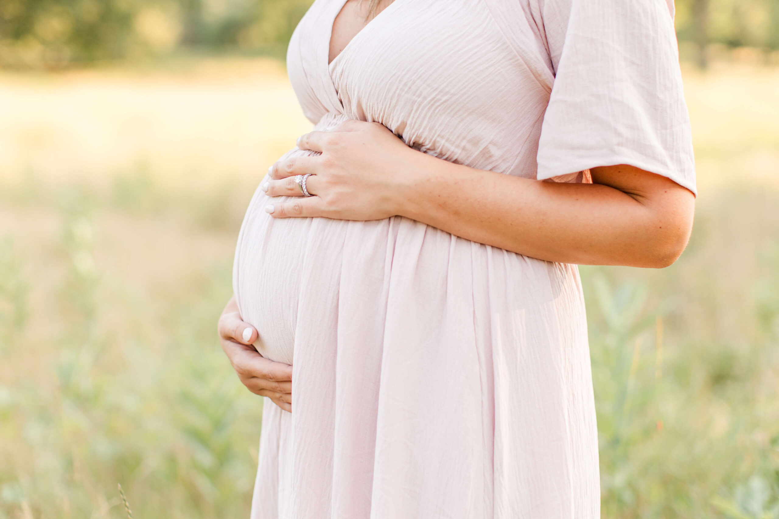 A close-up of a pregnant woman in a blush pink dress gently holding her baby bump with both hands, a ring visible on her finger