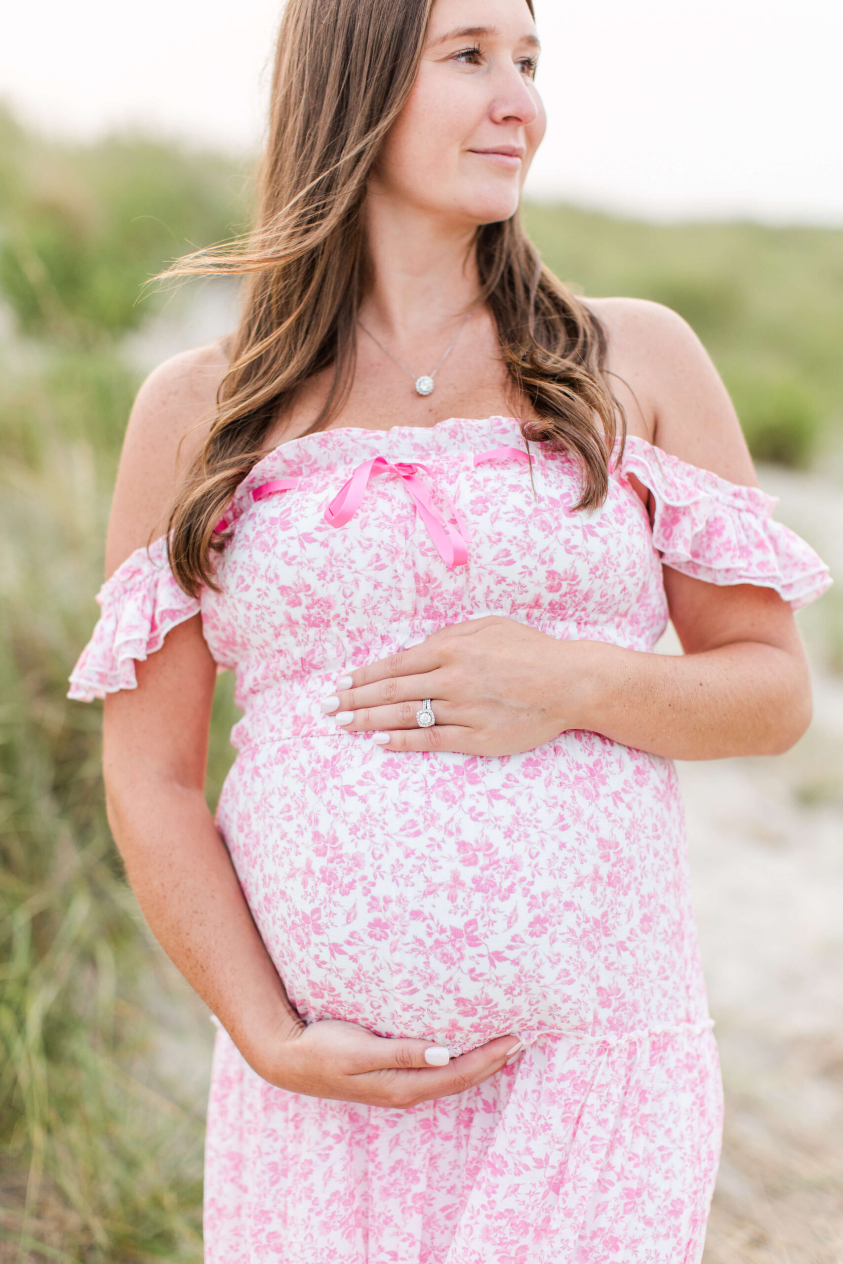 A close-up of a pregnant woman in a pink floral off-the-shoulder dress gently cradling her baby bump with both hands, a delicate necklace visible