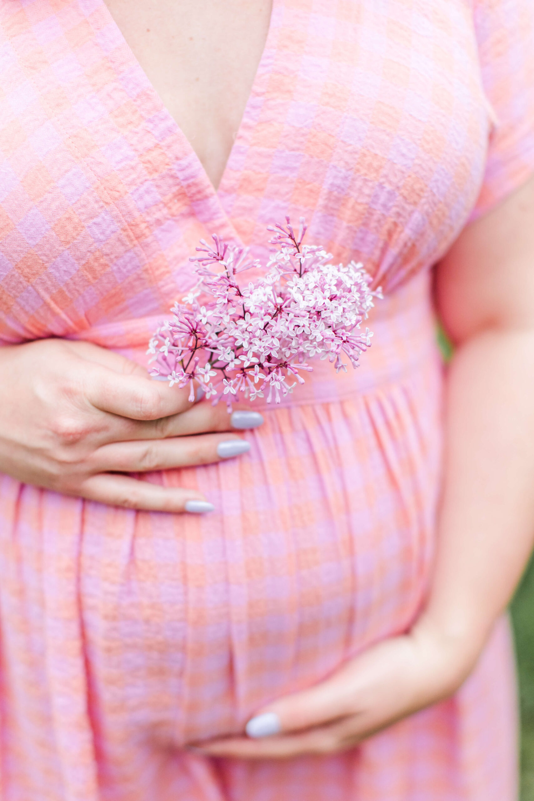 A close-up of a pregnant woman in a pink plaid dress holding a small bunch of pink lilac flowers against her baby bump