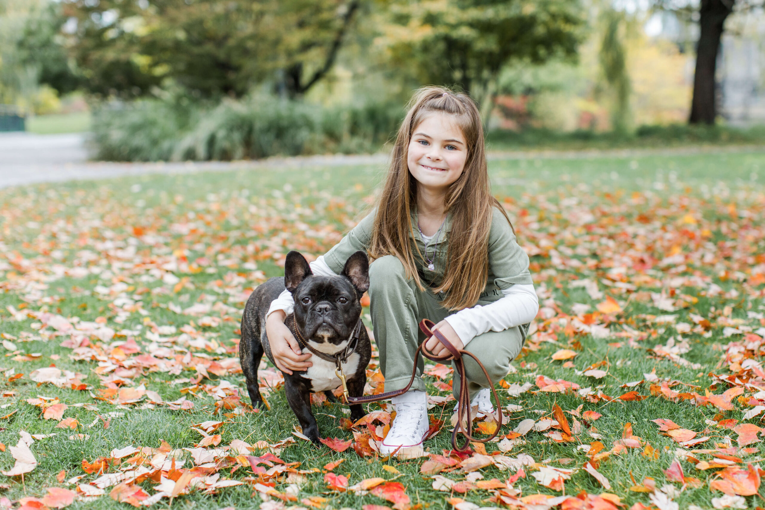 Young girl kneeling beside her black French bulldog during a dog photo shoot at North Point Park. They pose together on a lawn covered with fallen autumn leaves, with trees and park pathways visible in the background.