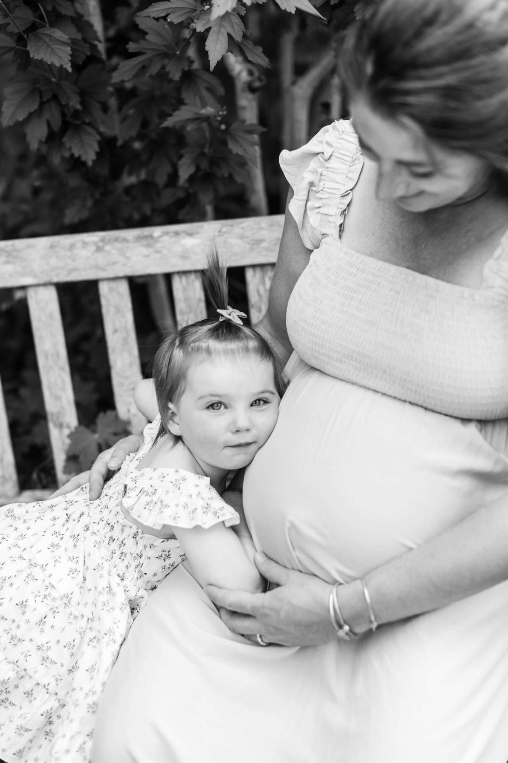 A black and white close-up of a toddler girl resting her head on her mother's pregnant belly while the mother looks down at her lovingly