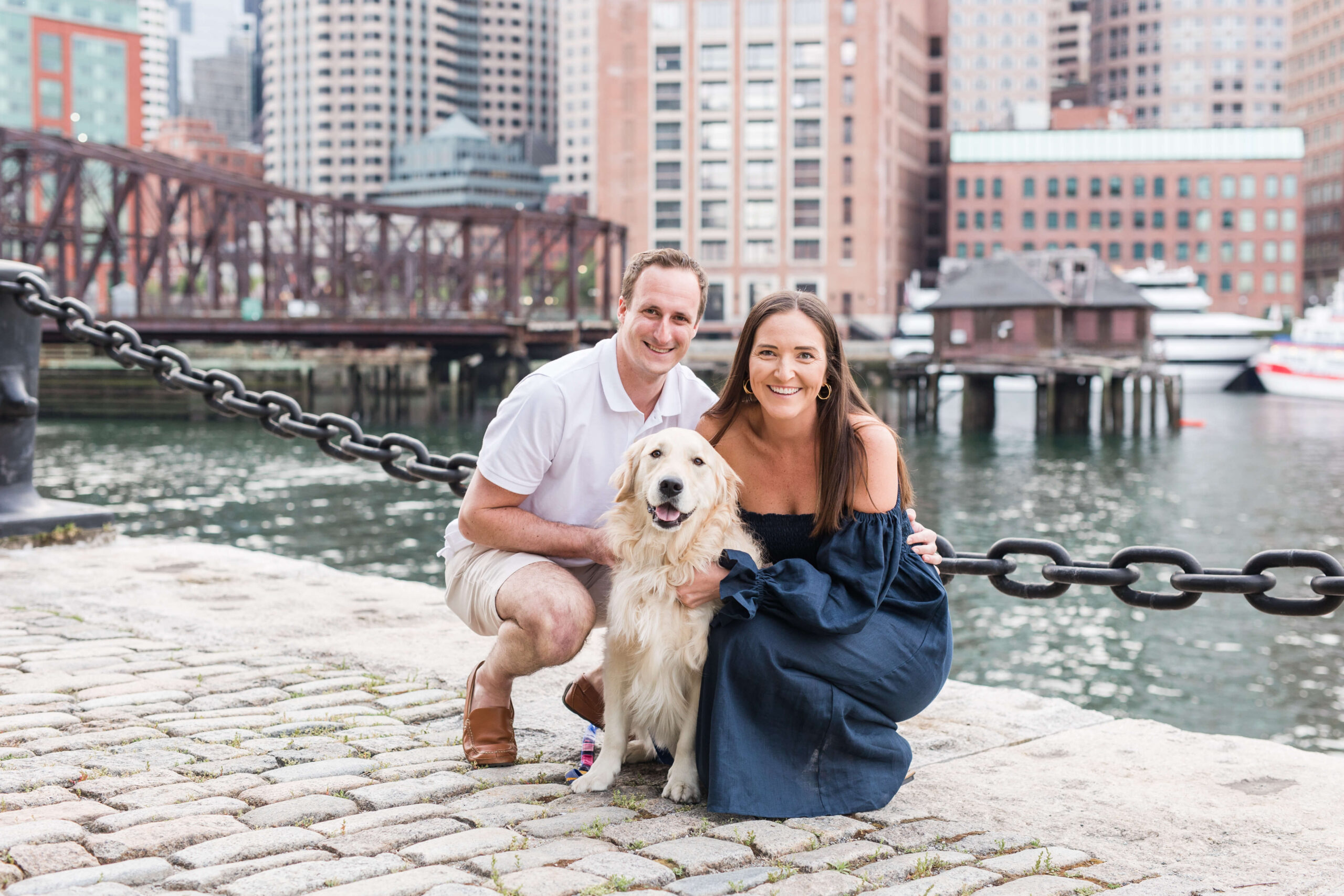 Couple kneeling beside their golden retriever during a dog photo shoot on a waterfront promenade. The city skyline and harbor create an urban backdrop behind the family portrait.