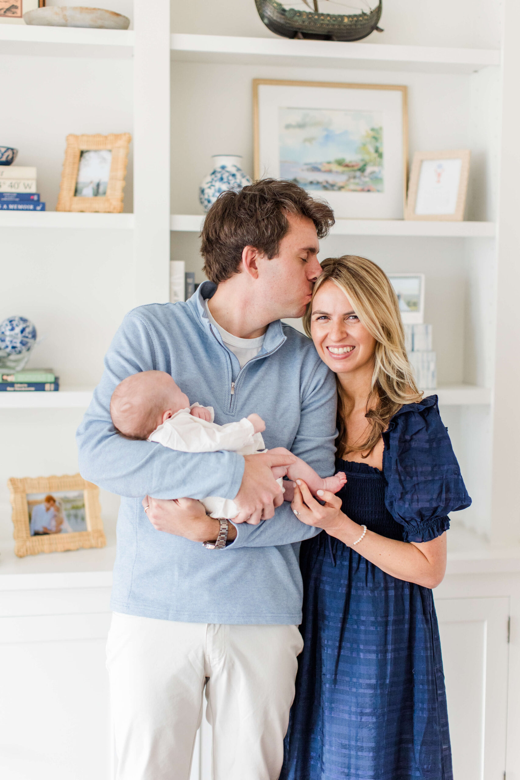 A couple in a cozy home with built-in white bookshelves, the man in a blue pullover kissing the woman's temple while they both hold their newborn baby