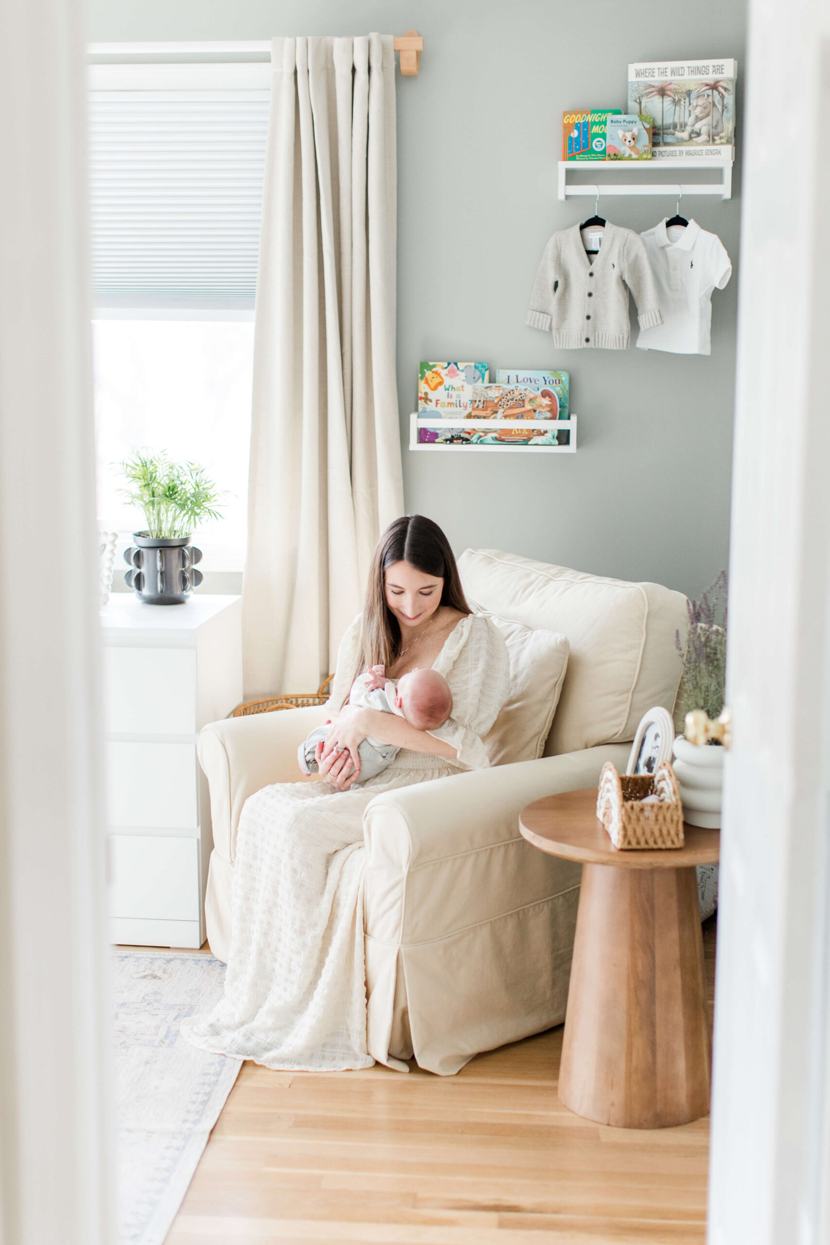 A mother sitting in a neutral-toned nursery chair by a window, gazing down lovingly at her newborn baby wrapped in a white blanket